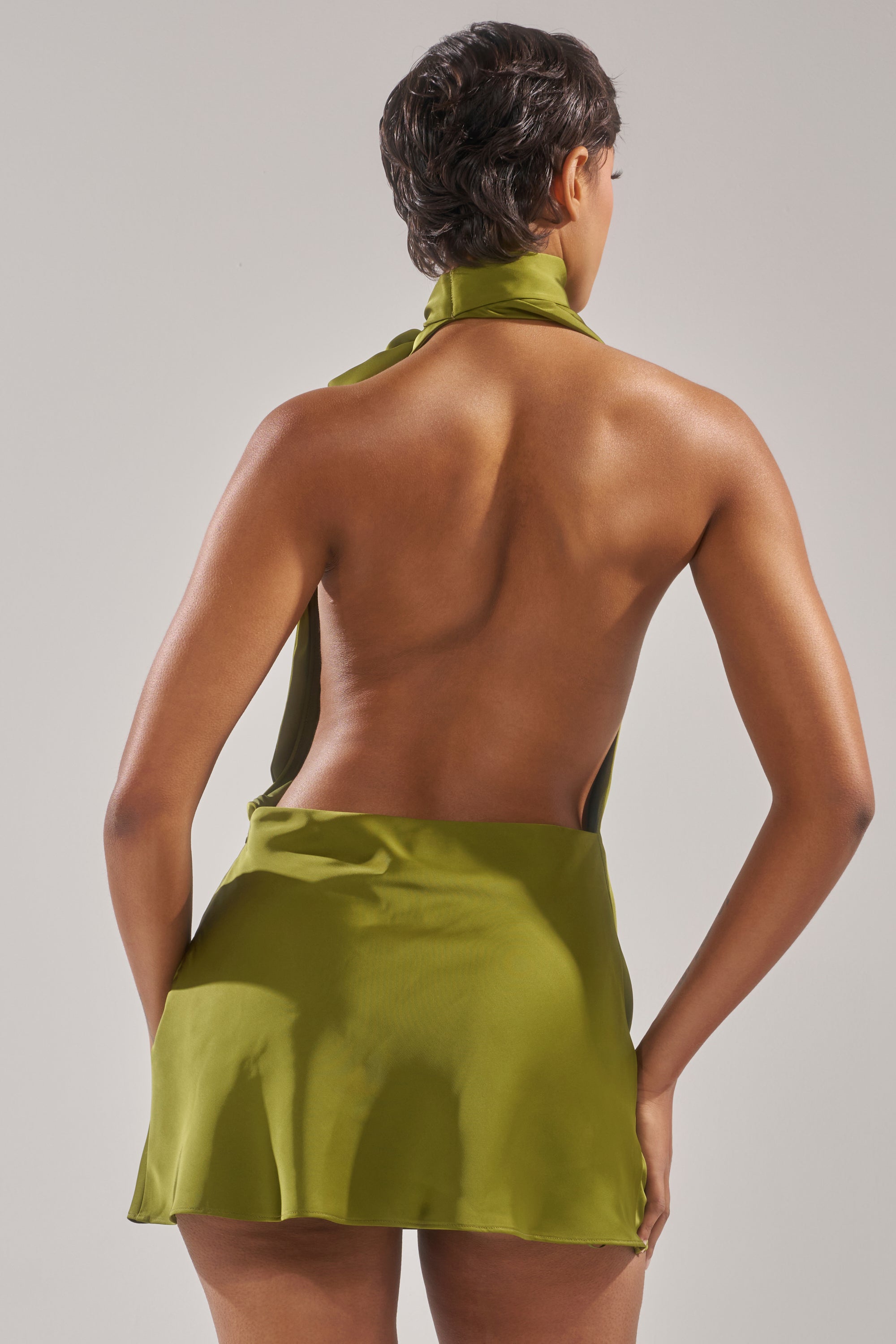 A person with short dark hair faces away, wearing the GIGI MINI DRESS IN GREEN—an olive halter cowl neck mini dress that bares their back and upper thighs. The backdrop is plain and light.