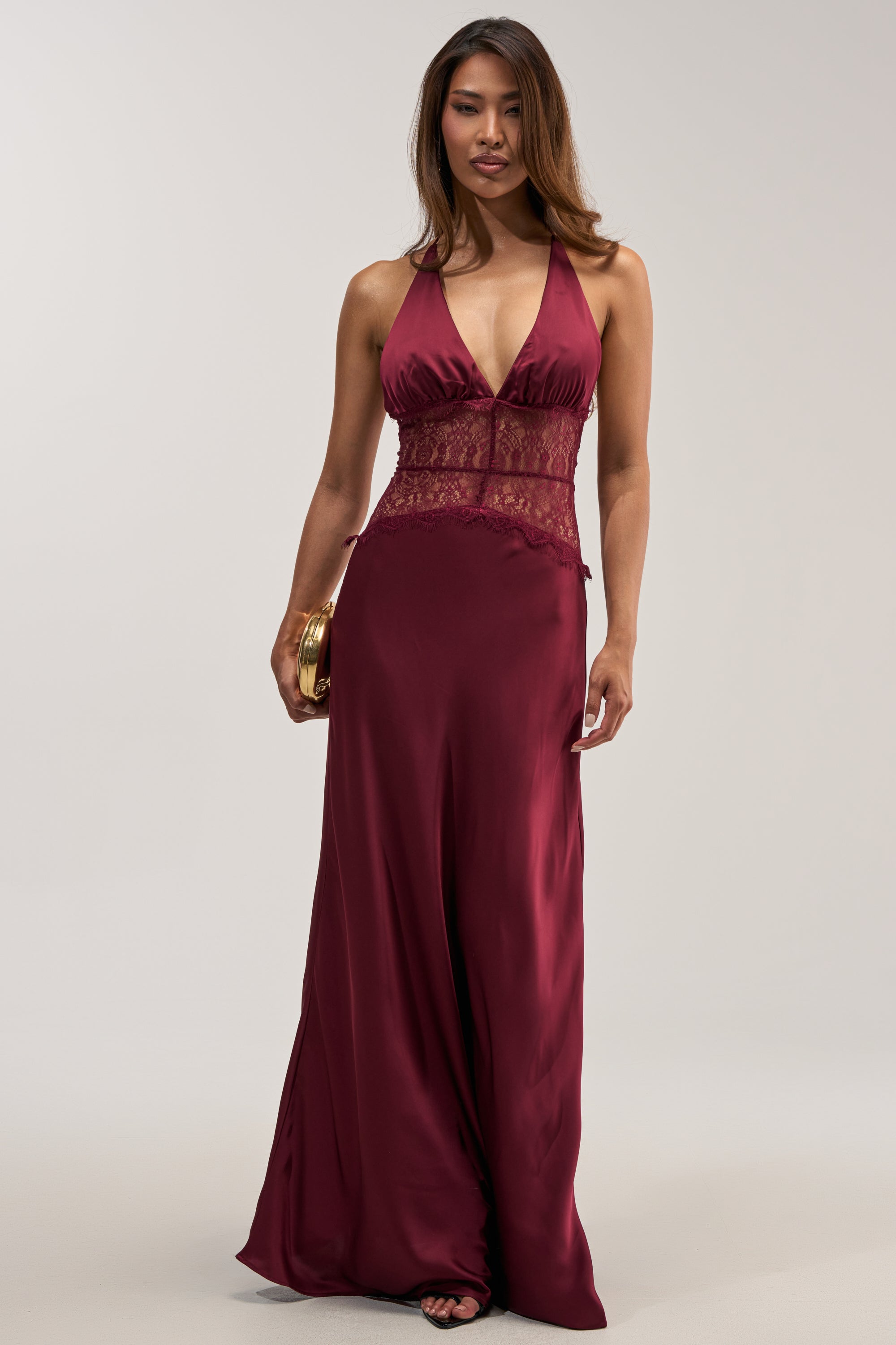 A woman wearing the LUXE LIFE SATIN LACE MAXI DRESS—a sleeveless, deep red satin gown featuring delicate lace accents at the waist—holds a small gold clutch as she stands against a plain light background.