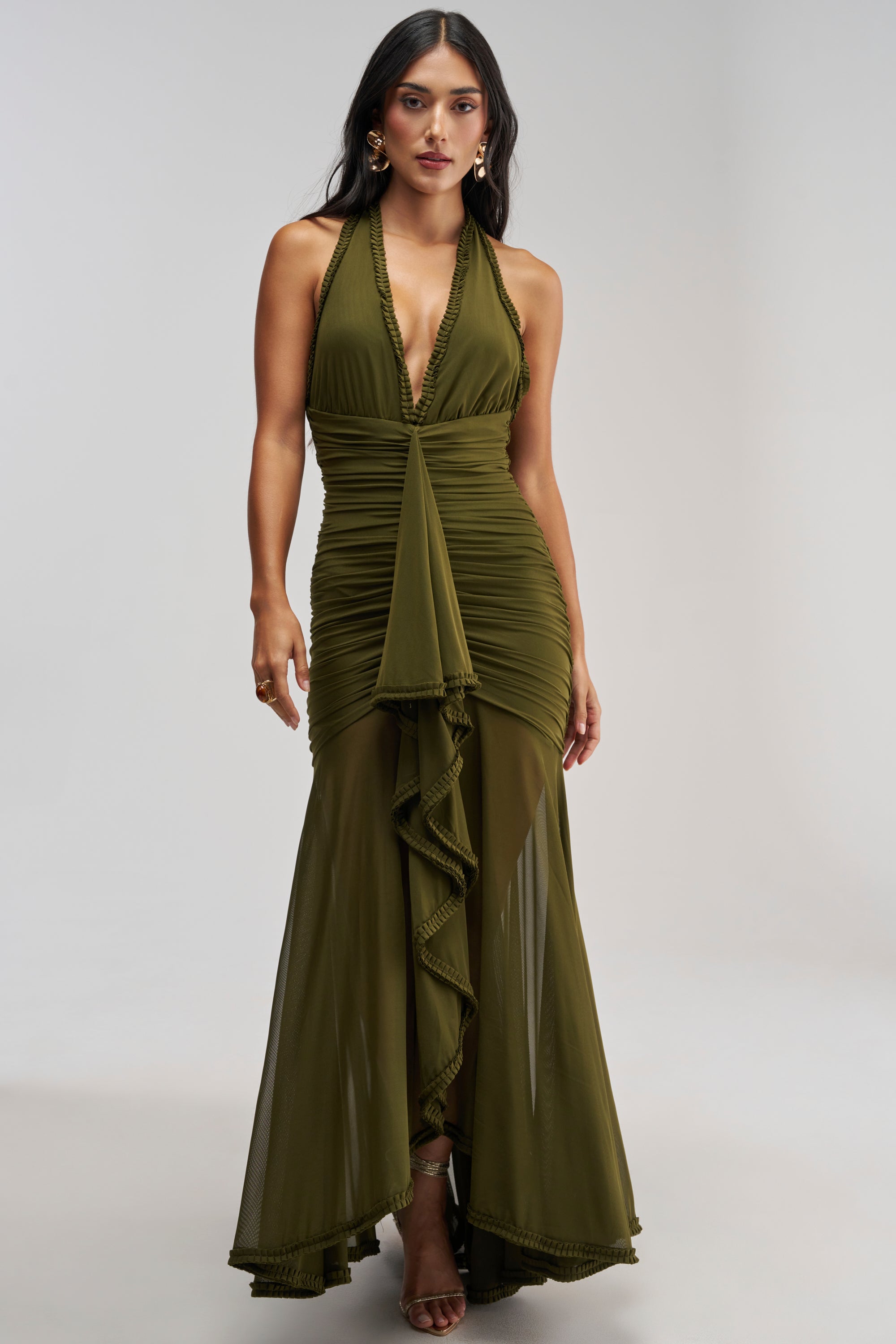 A woman wearing the SWEET SUNSET MESH MAXI DRESS IN OLIVE, a sleeveless halter-neck evening gown with a ruched bodice, ruffled front detail, and sheer skirt panels, stands against a plain light gray background. This stunning olive mesh maxi dress makes an elegant choice for a wedding guest dress.
