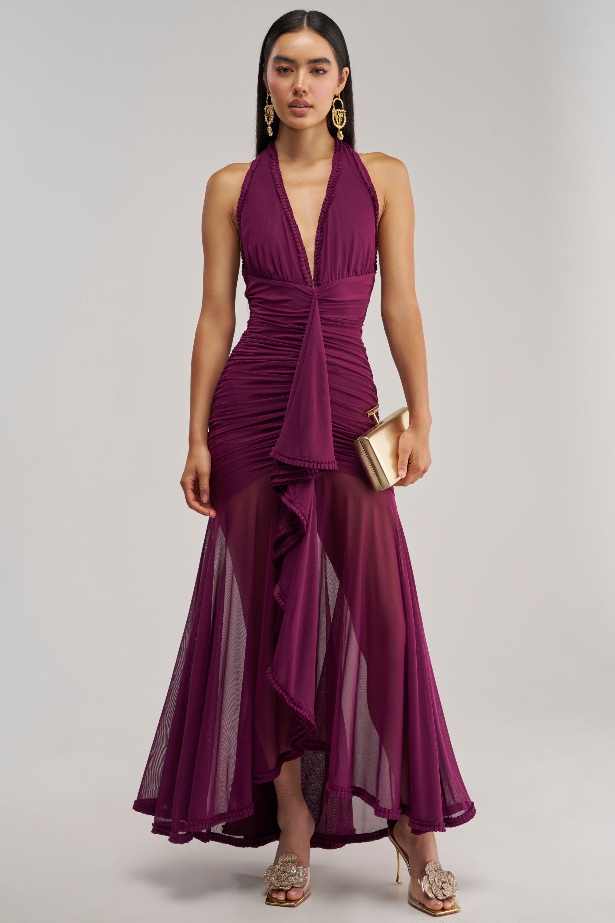 Wearing the SWEET SUNSET MESH MAXI DRESS IN PLUM—a sleeveless maxi dress featuring ruffled detailing and a chic ruffle hem—a woman completes her look with a gold clutch, statement earrings, and heeled sandals, creating the perfect stylish ensemble for a wedding guest.