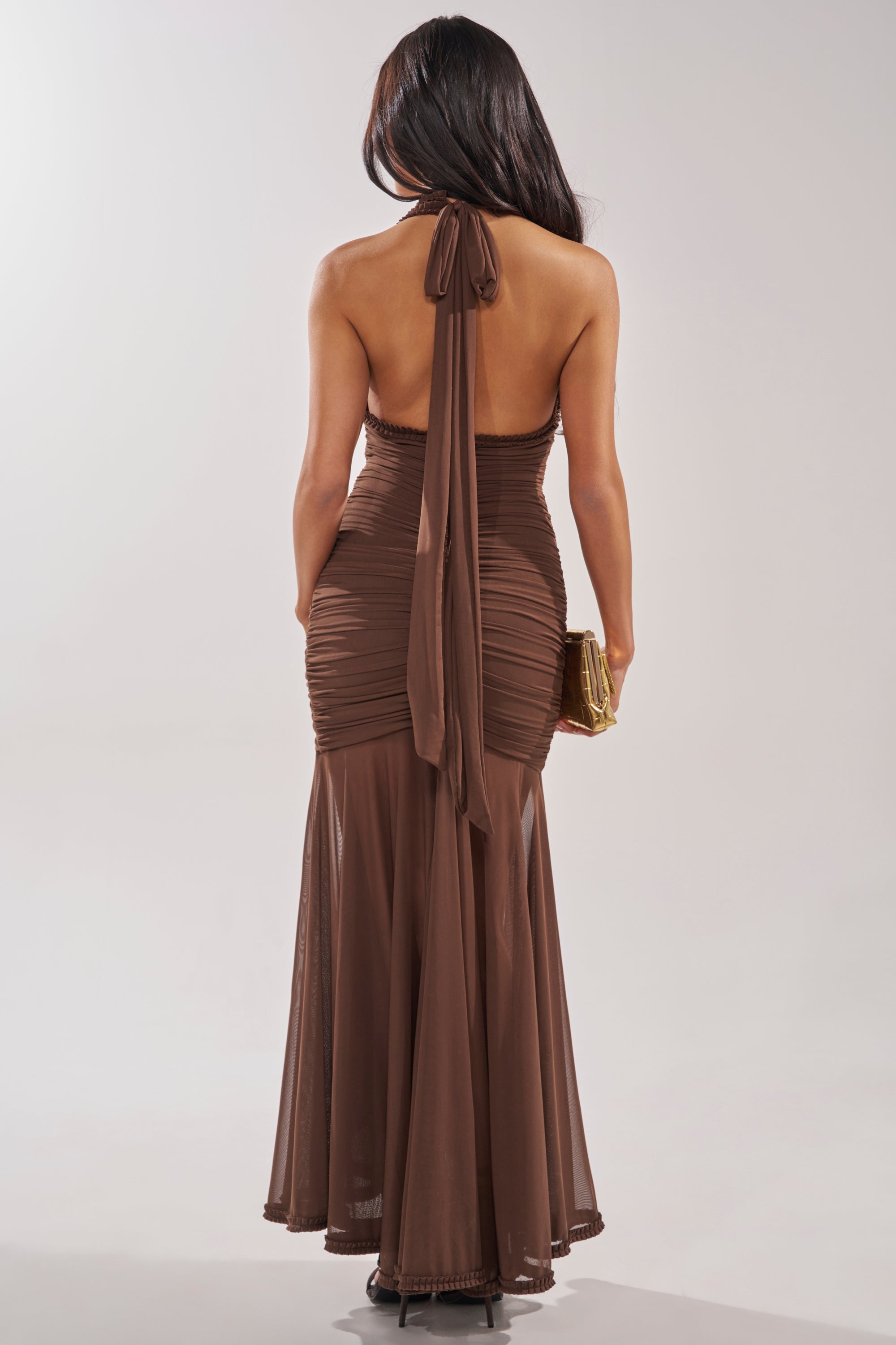 A woman with long dark hair is seen from behind wearing the SWEET SUNSET MESH MAXI DRESS, a ruched backless mermaid gown with a halter-neck and long tie, holding a small gold clutch against a neutral background. 4