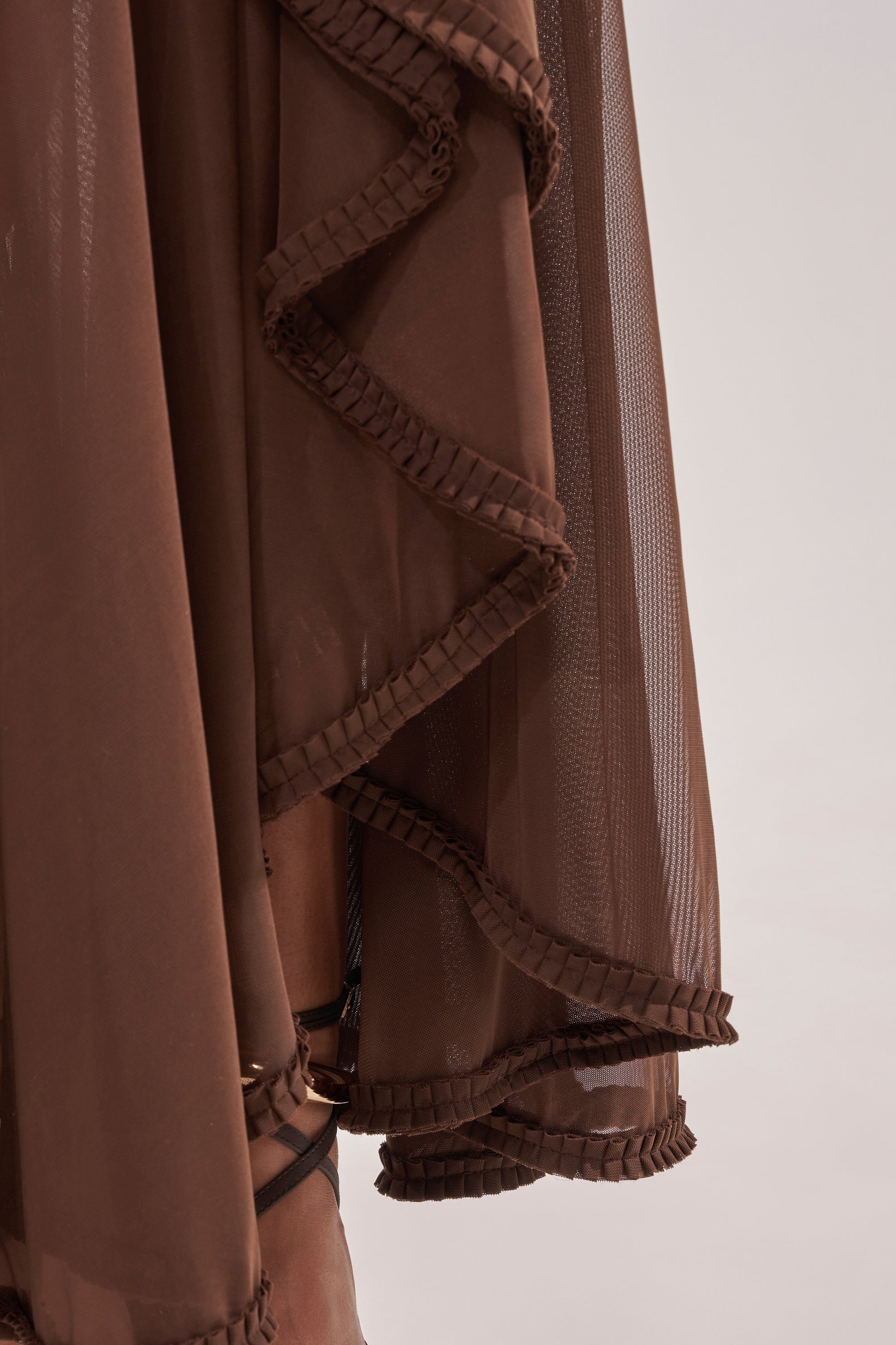 A close-up of the SWEET SUNSET MESH MAXI DRESS highlights its sheer brown halter style, layered ruffled skirt, and lightweight, semi-transparent mesh fabric elegantly draping over the legs. 7
