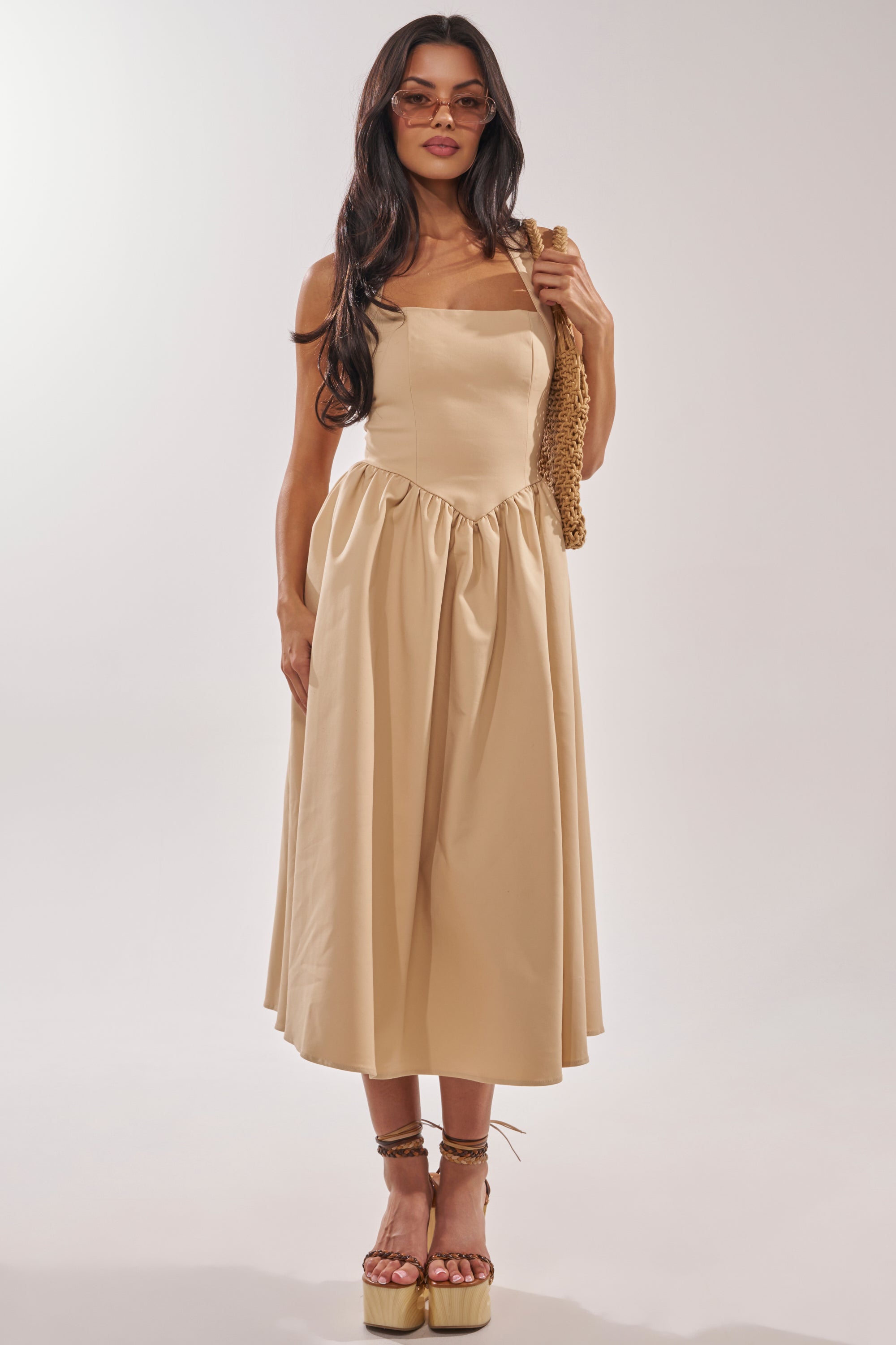A woman wears the SWEET SOFIE HALTER MIDI DRESS, featuring a khaki corset-inspired bodice and gathered skirt. She accessorizes with sunglasses, wedge sandals, and a woven bag over her shoulder against a light background. 0