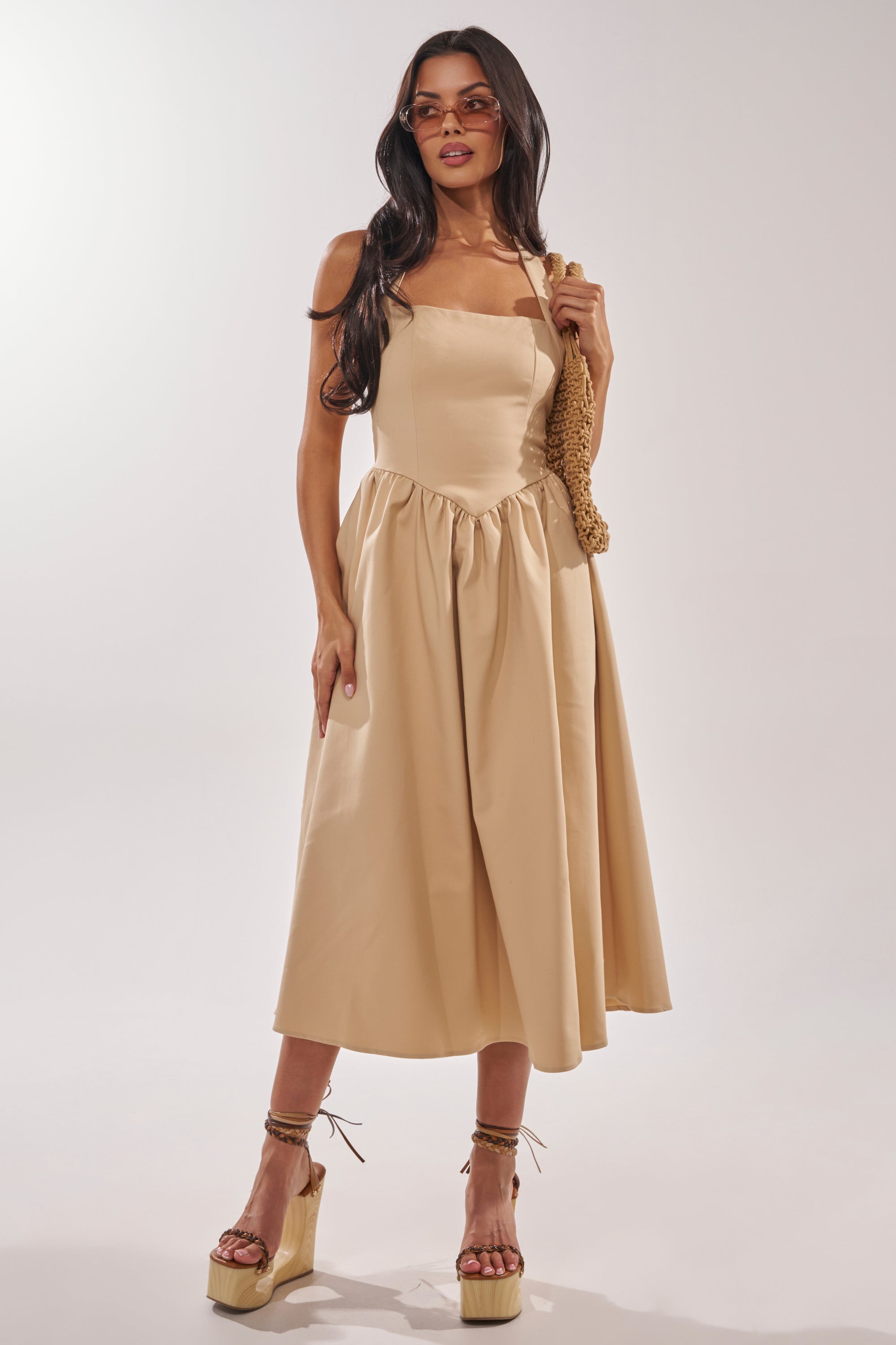 A woman in the SWEET SOFIE HALTER MIDI DRESS and platform sandals stands against a plain background, holding a textured shoulder bag. She has long, wavy dark hair and wears sunglasses. 2