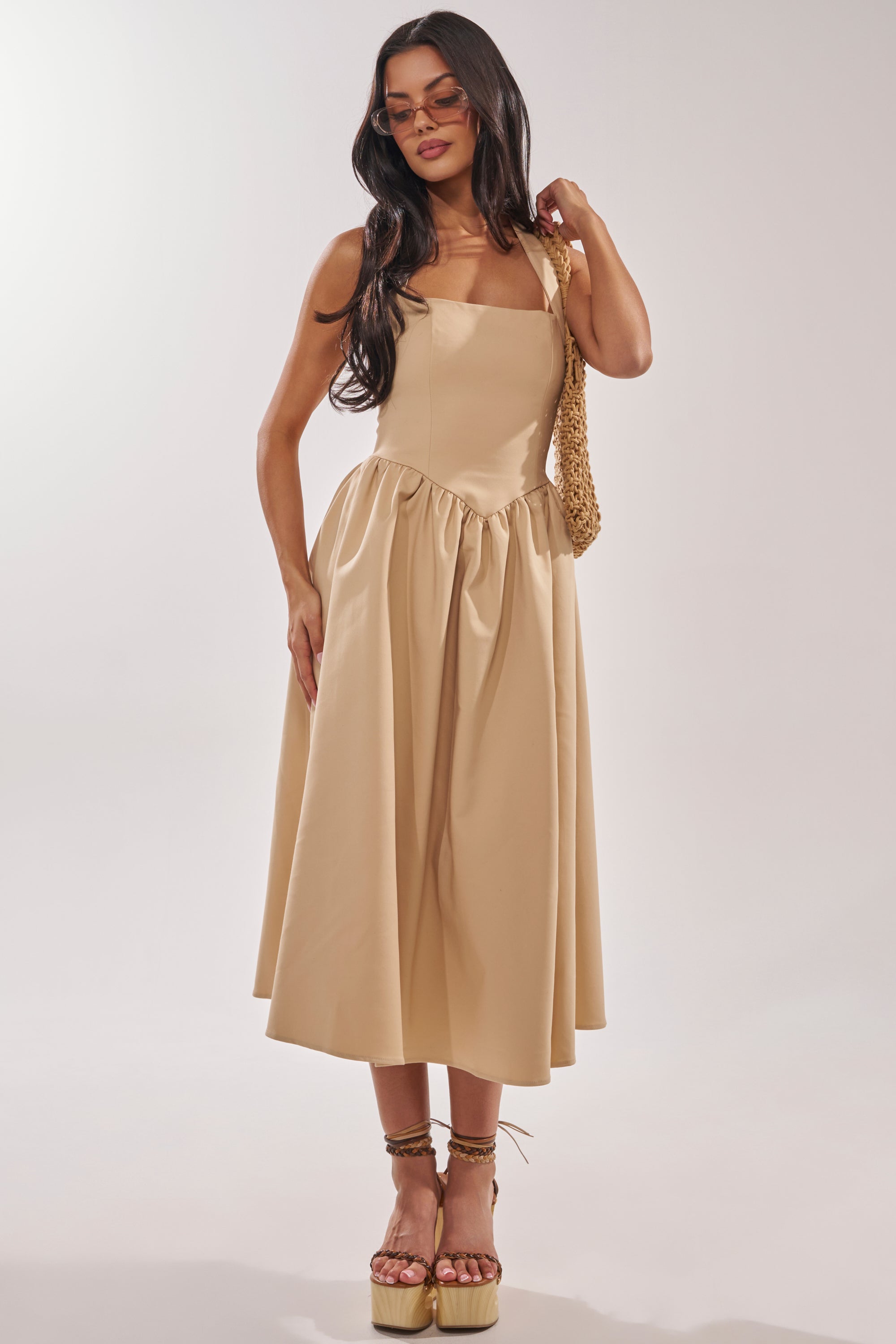 A woman with long dark hair wears the SWEET SOFIE HALTER MIDI DRESS, featuring a corset-inspired bodice and flared skirt. She pairs it with sunglasses, platform sandals, and a woven shoulder bag against a plain light background. 3