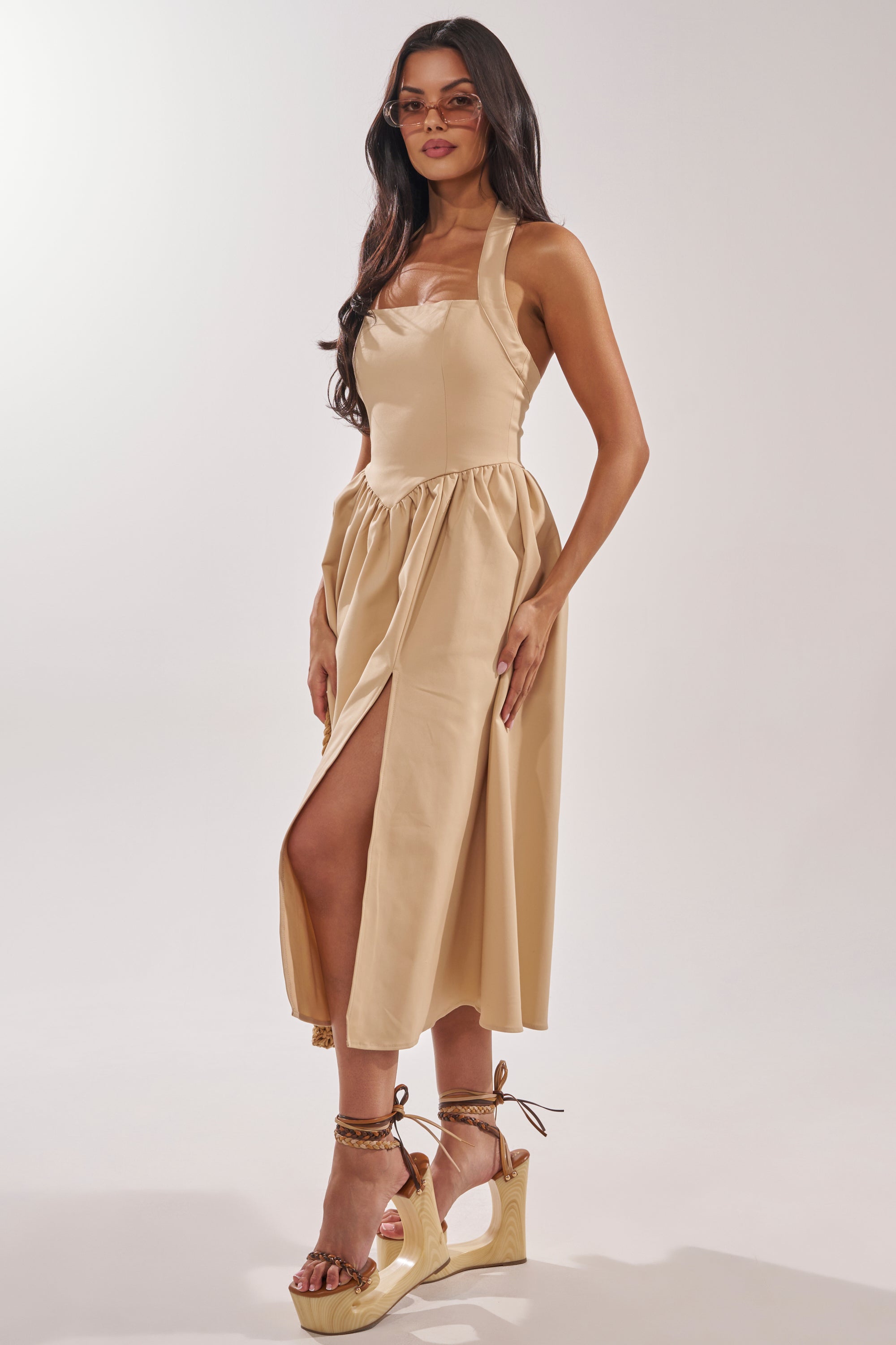 A woman poses against a plain light background wearing the SWEET SOFIE HALTER MIDI DRESS in khaki, paired with tan wedge sandals and sunglasses. 4