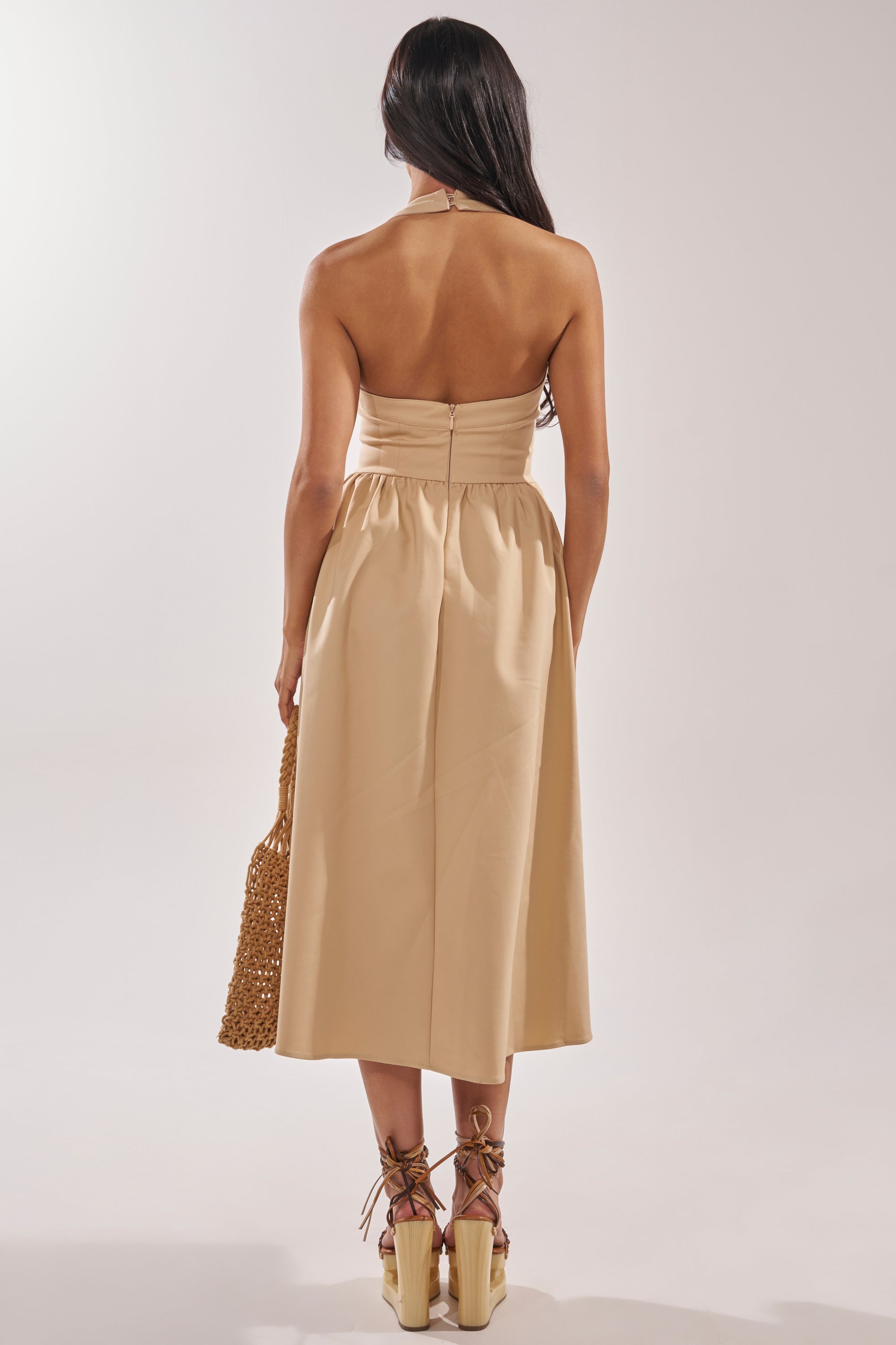 A woman with long dark hair wears the SWEET SOFIE HALTER MIDI DRESS in khaki and platform sandals, holding a woven bag in her left hand. She faces away against a plain light gray background. 5