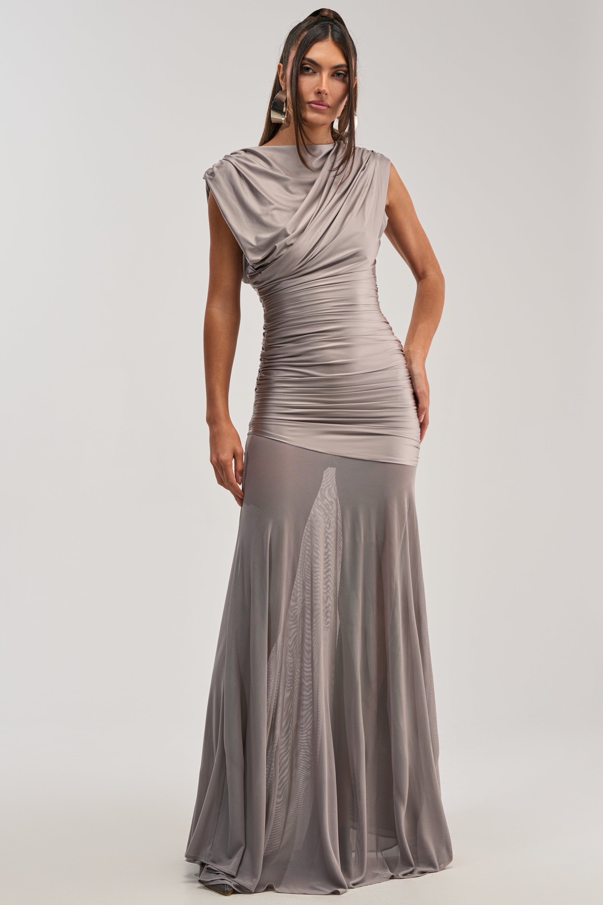 A woman wears the LIMITLESS SATIN MAXI DRESS, a floor-length taupe gown with ruched details, draped sleeves, and a sheer flared skirt. Fitted at the bodice and hips, she stands against a plain light background.