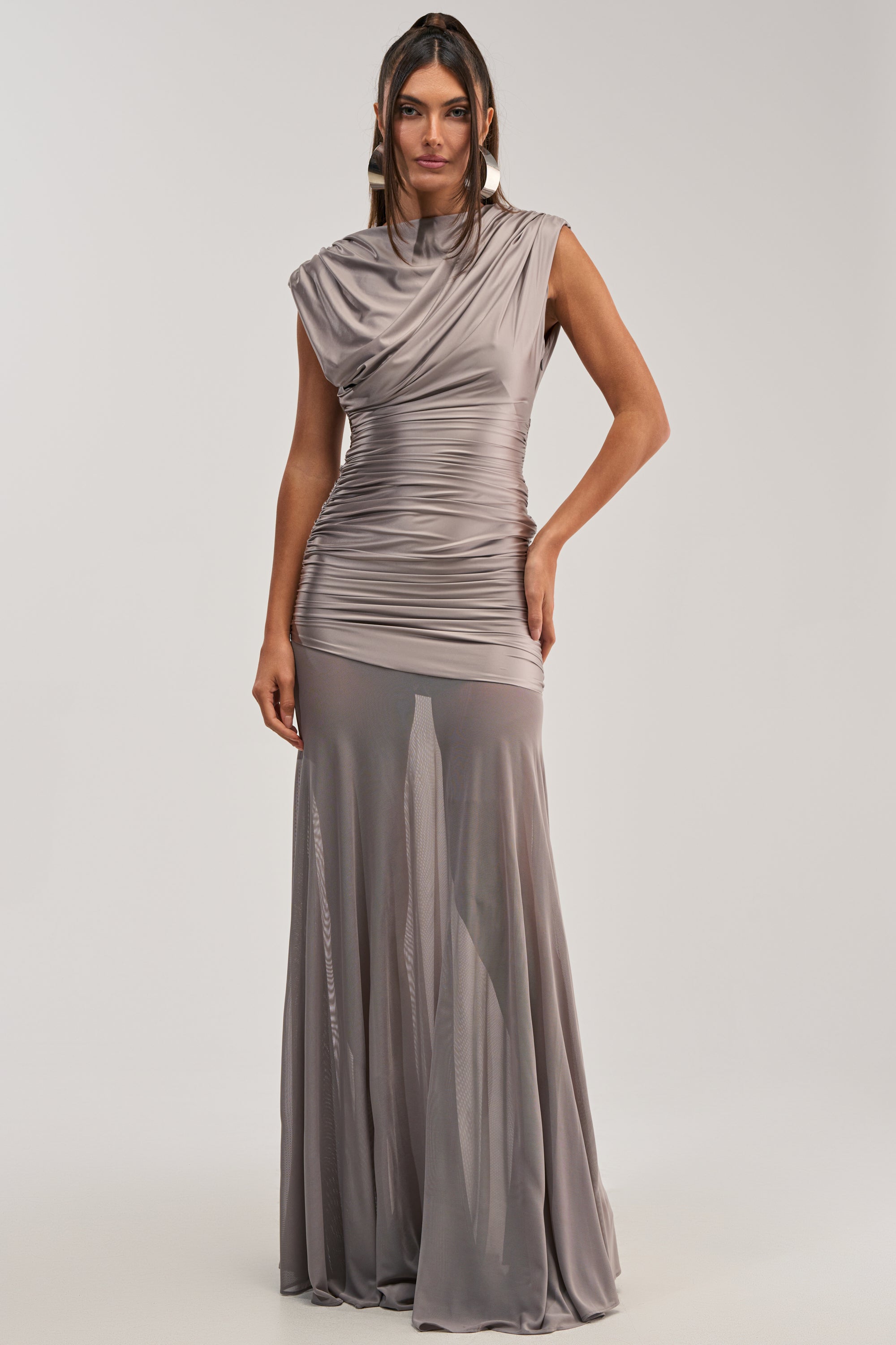 A woman poses against a plain background in the LIMITLESS SATIN MAXI DRESS—a silver, sleeveless satin gown with ruched details, a draped high neckline, and a slightly sheer floor-length skirt—ideal for formal occasions.