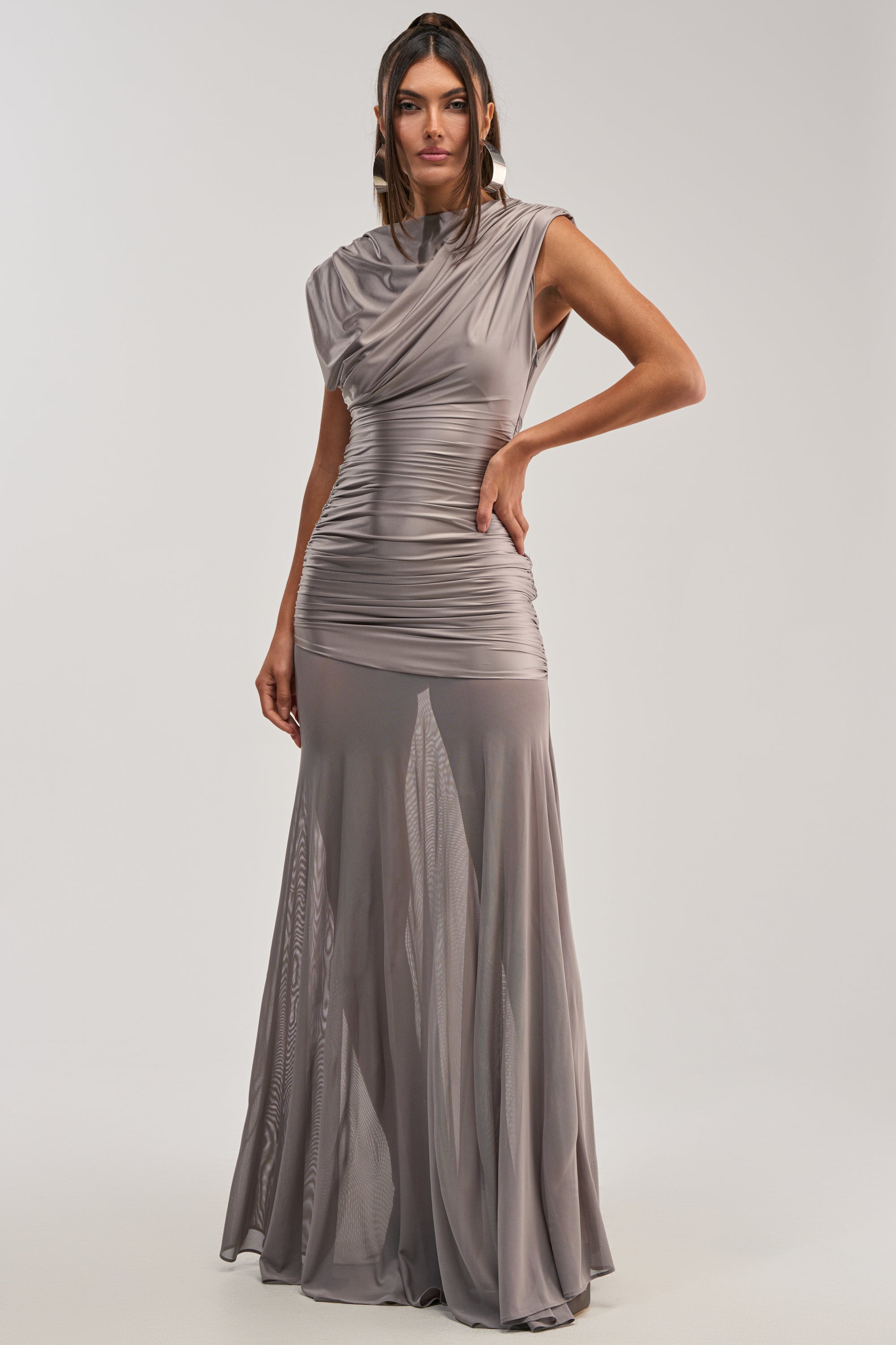 A woman stands against a plain light background wearing the LIMITLESS SATIN MAXI DRESS, a sleeveless, floor-length silver gown featuring a fitted ruched bodice and flowing skirt—an elegant choice for formal events.