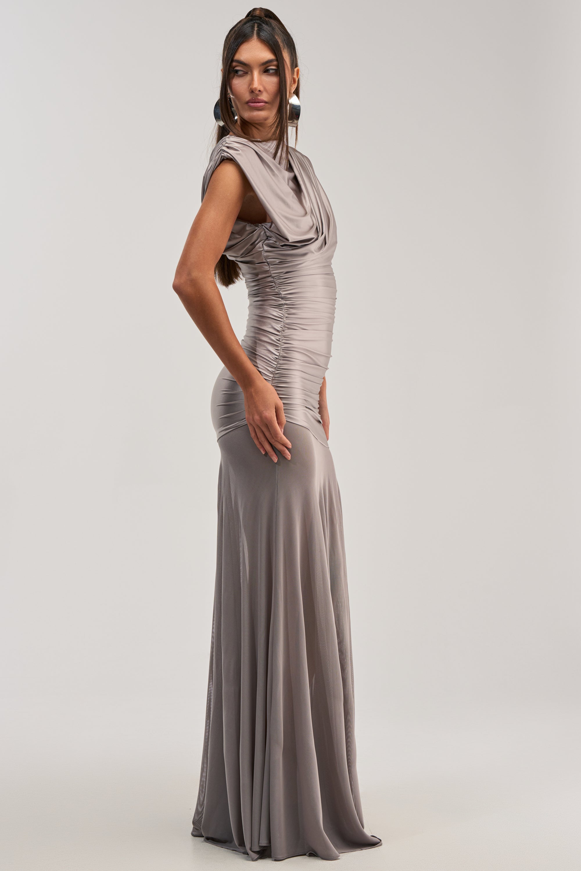 Woman in profile wears the LIMITLESS SATIN MAXI DRESS, a long silver fitted gown with ruched detailing. Paired with large circular earrings and straight dark hair, it’s an elegant look for formal events.