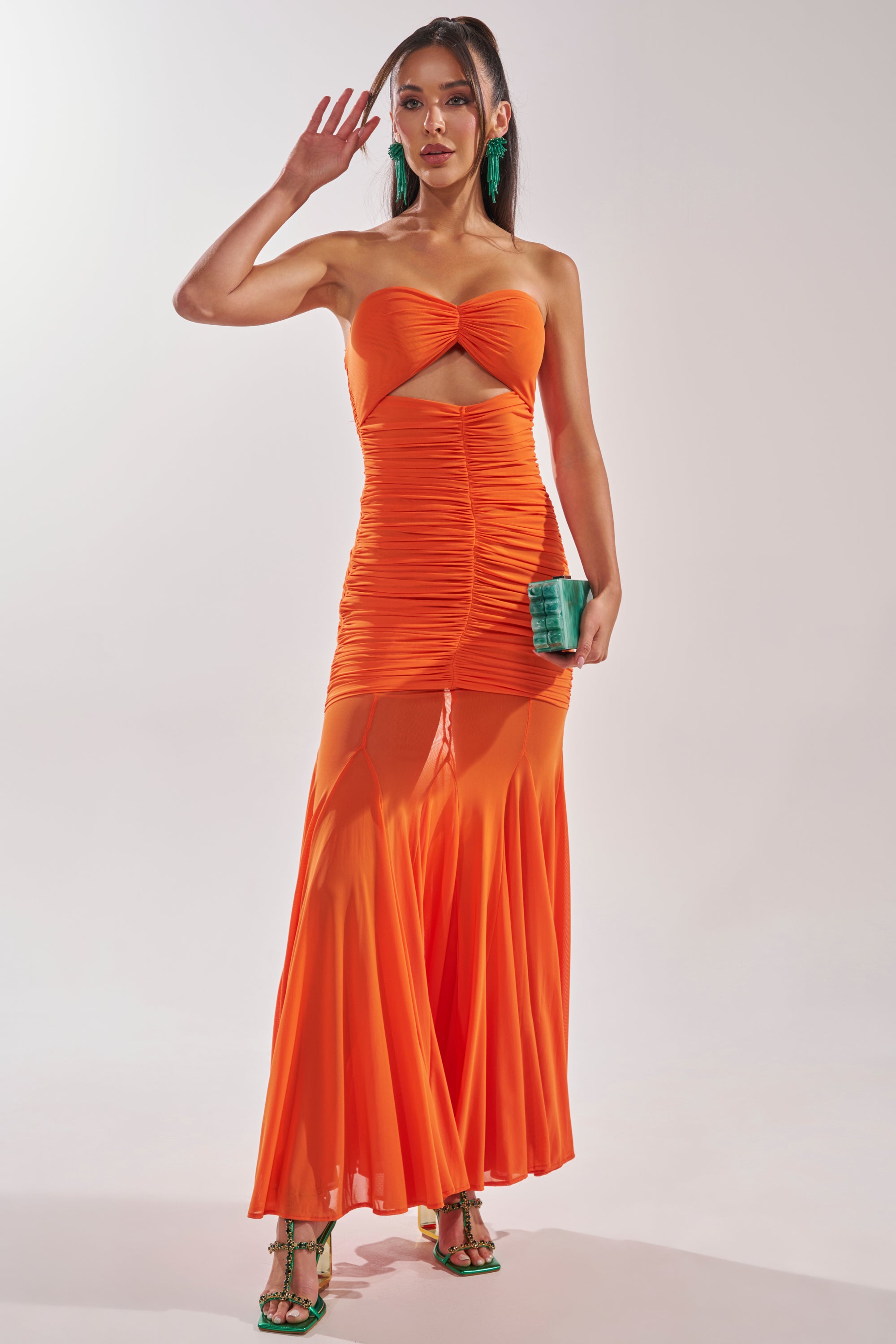 A woman wears the CABANA CHILL CINCHED MAXI DRESS, a strapless ruched orange maxi with a front cutout and sheer skirt. She styles it with green heels, a matching clutch, pigtails, and statement earrings for an evening occasion. 1