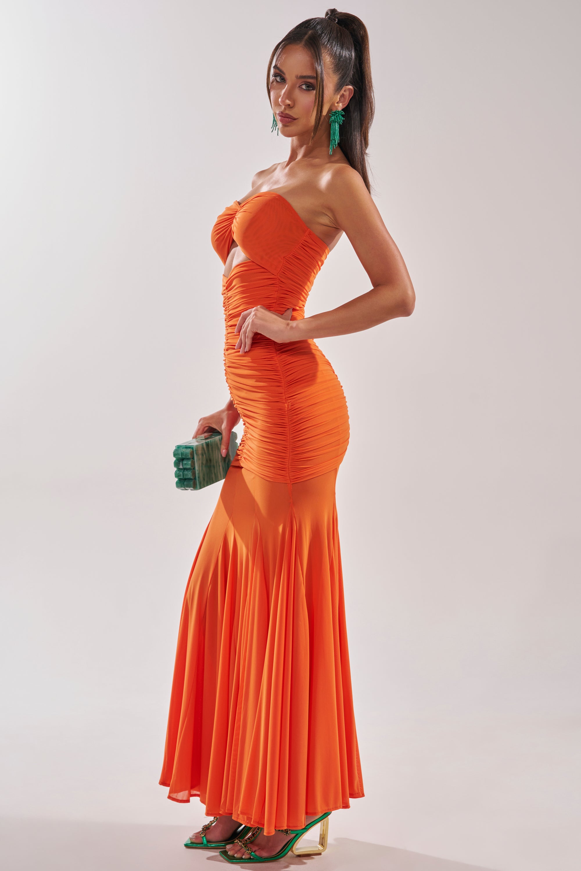 A woman poses sideways in the CABANA CHILL CINCHED MAXI DRESS, a bright orange strapless ruched maxi. She carries a green clutch, wears matching green heels and statement earrings, with her long hair in a high ponytail against a light background. 3