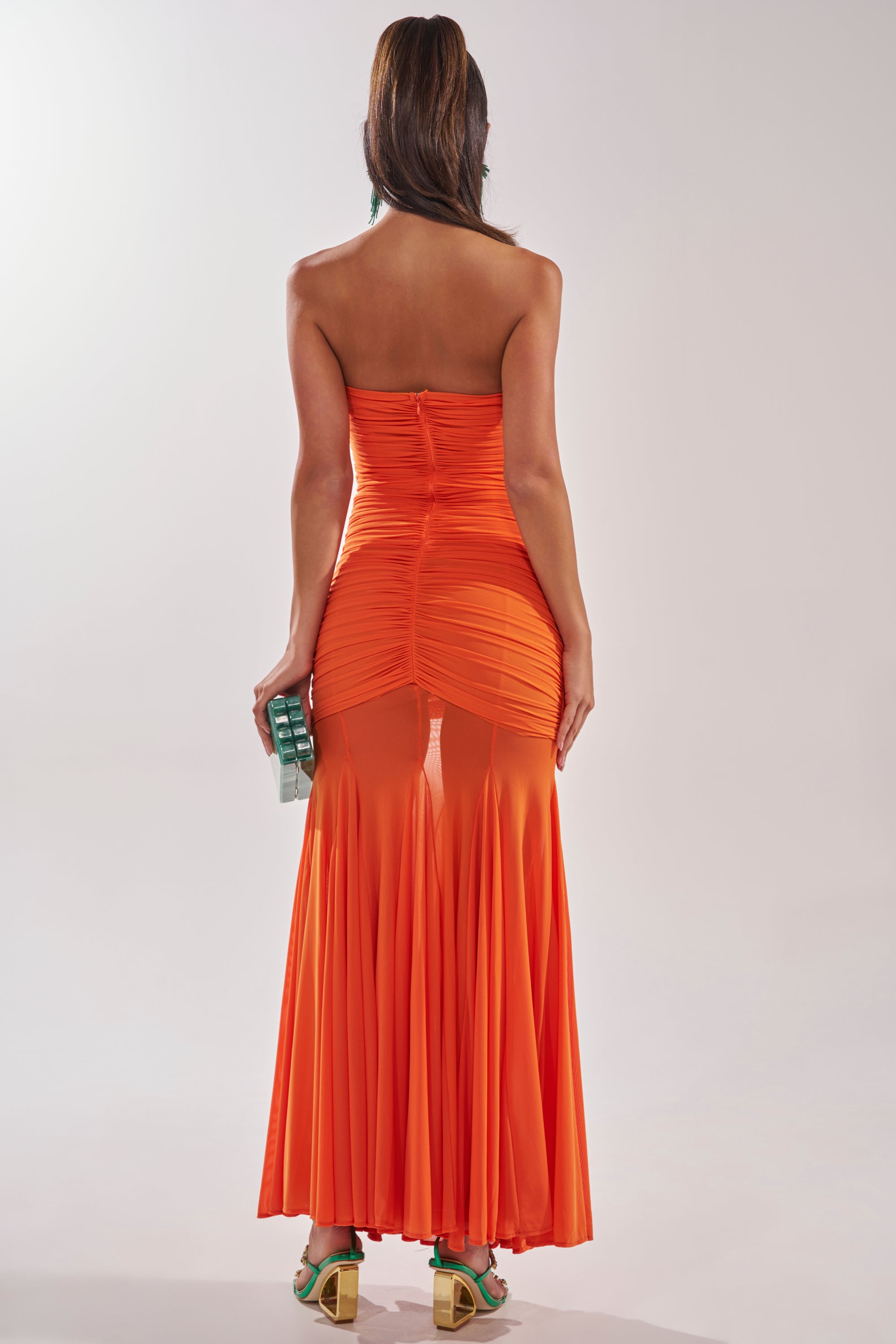A woman faces away, wearing the CABANA CHILL CINCHED MAXI DRESS—an orange strapless maxi with ruched details, sheer panels, and a flared hem. She holds a teal clutch and sports green heels against a plain light background. 4