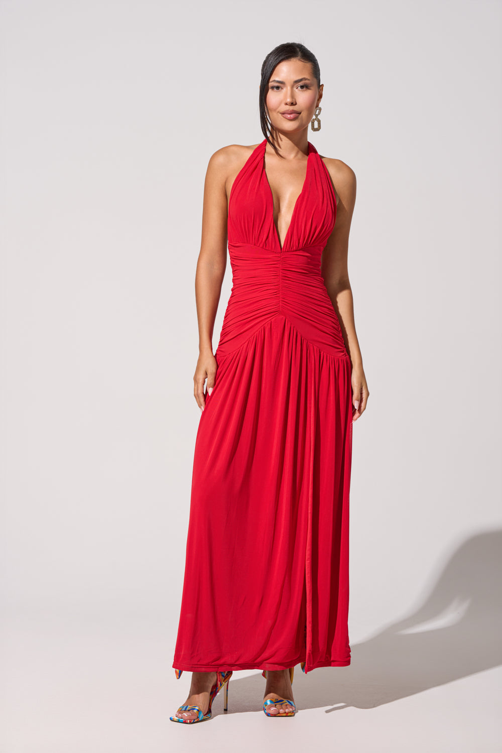 A woman with straight dark hair and hoop earrings wears the SUGAR RUSH MESH MAXI DRESS, a red halter maxi with a plunge neckline and ruched details, standing against a plain background in colorful high-heeled shoes. 1