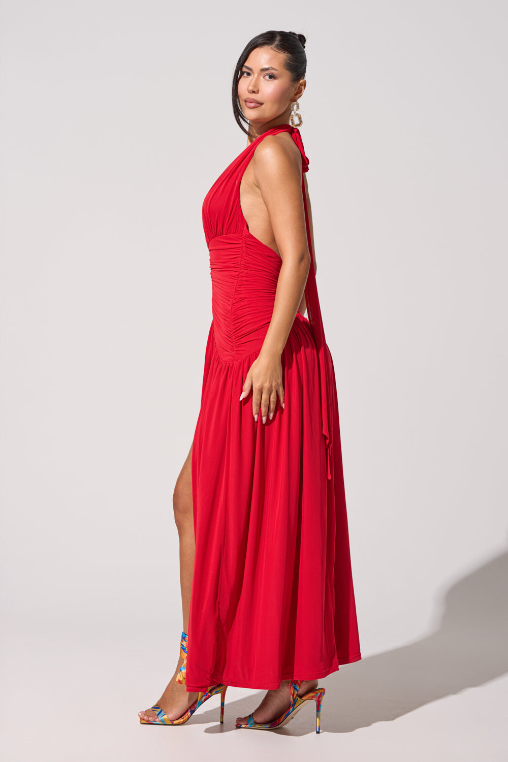 A woman stands sideways against a light background wearing the SUGAR RUSH MESH MAXI DRESS, a ruched red halter maxi with a high slit, colorful heels, her hair tied back, and statement earrings completing the look. 3