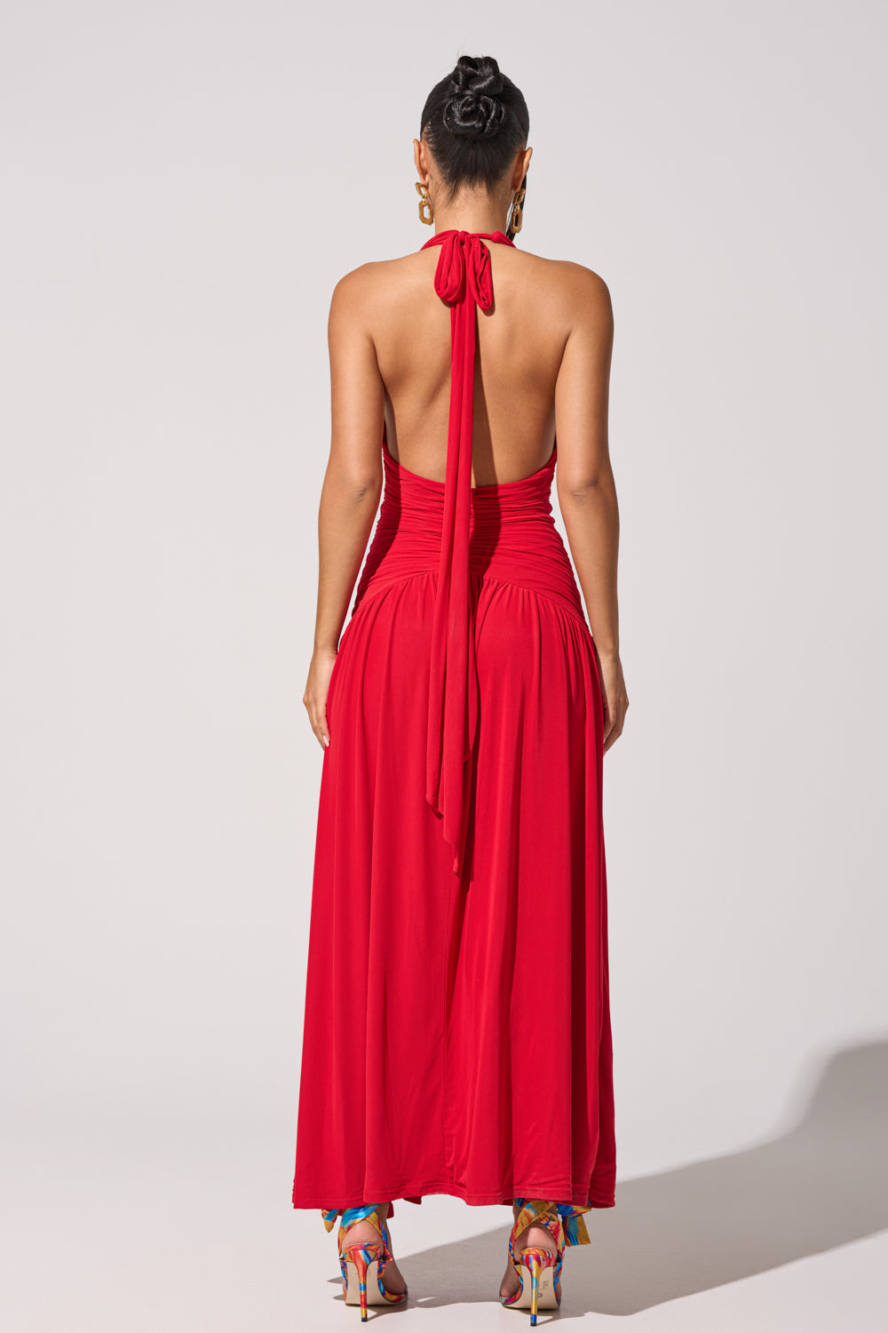 A woman faces away from the camera in the SUGAR RUSH MESH MAXI DRESS, a ruched red halter gown with a neck bow. She completes her look with colorful heels, gold hoops, and a sleek bun. 4