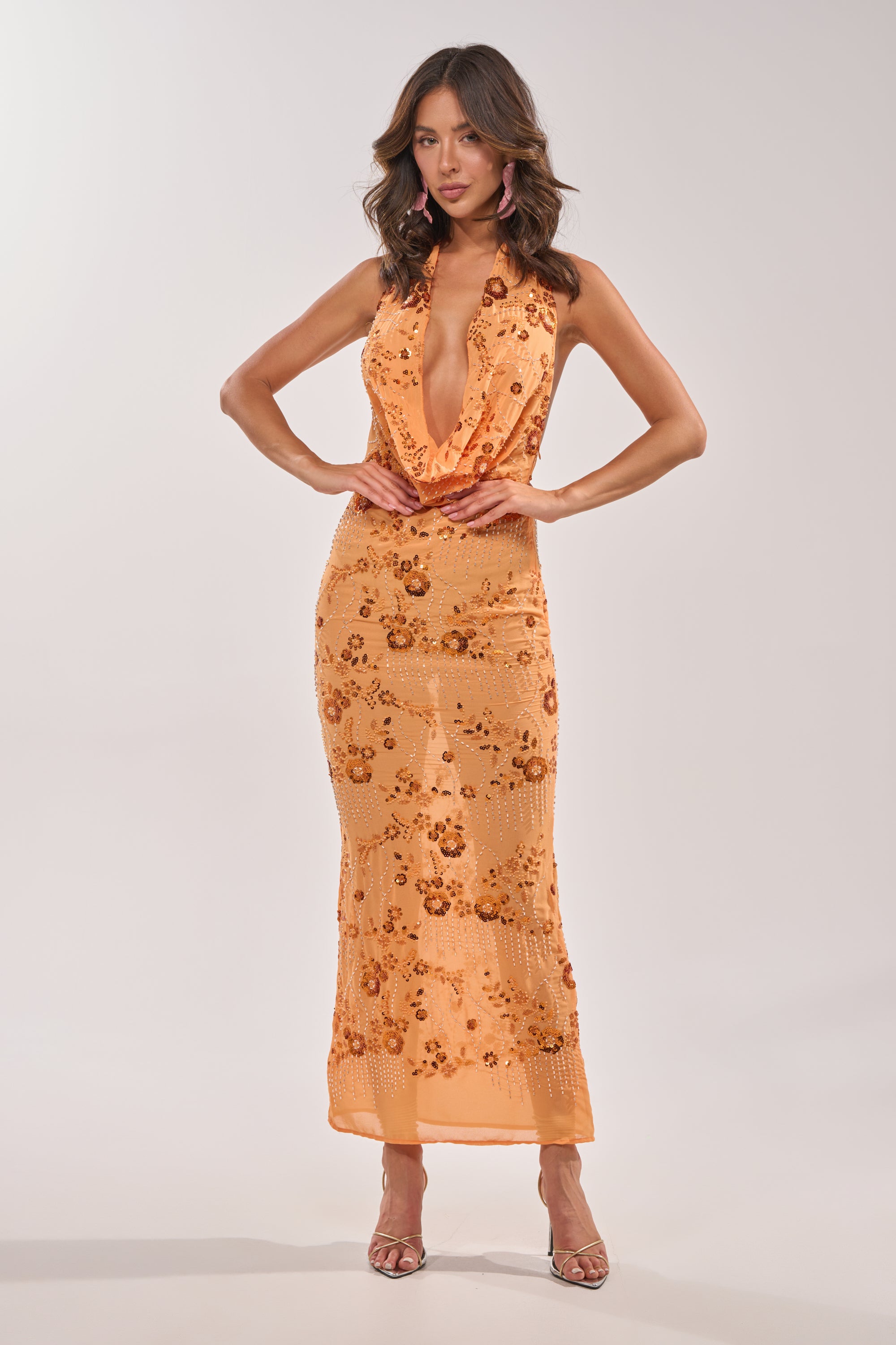 In a studio, a woman with wavy hair and hands on hips models the LOVE LETTERS EMBELLISHED MAXI DRESS IN ORANGE—an orange beaded halter maxi dress with sequin floral designs, a plunging neckline, and strappy heeled sandals. 2