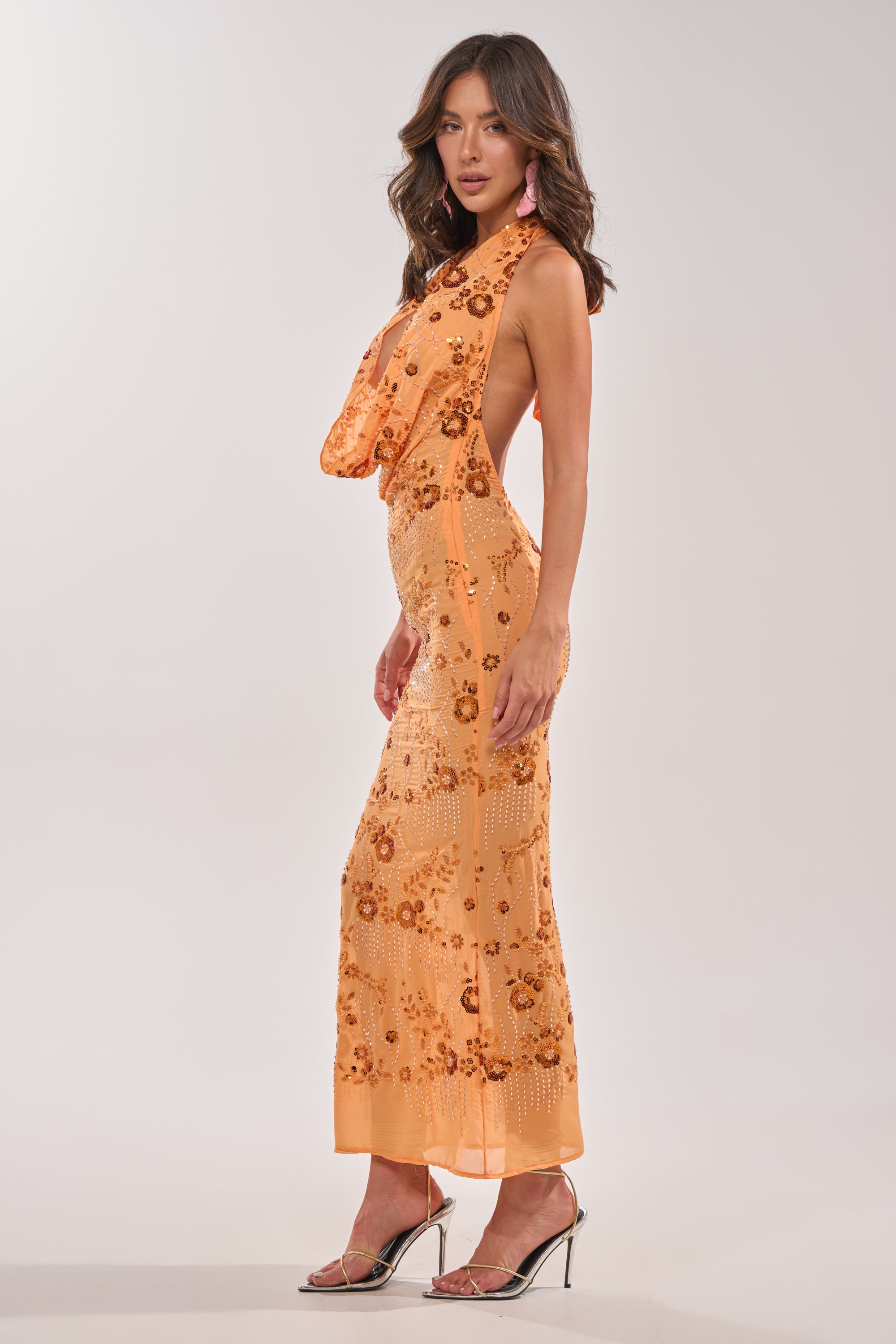 A woman poses in the LOVE LETTERS EMBELLISHED MAXI DRESS IN ORANGE—a floor-length gown with sequin floral patterns and an open back. She pairs it with wavy hair, pink earrings, and silver high-heeled sandals against a plain background. 4