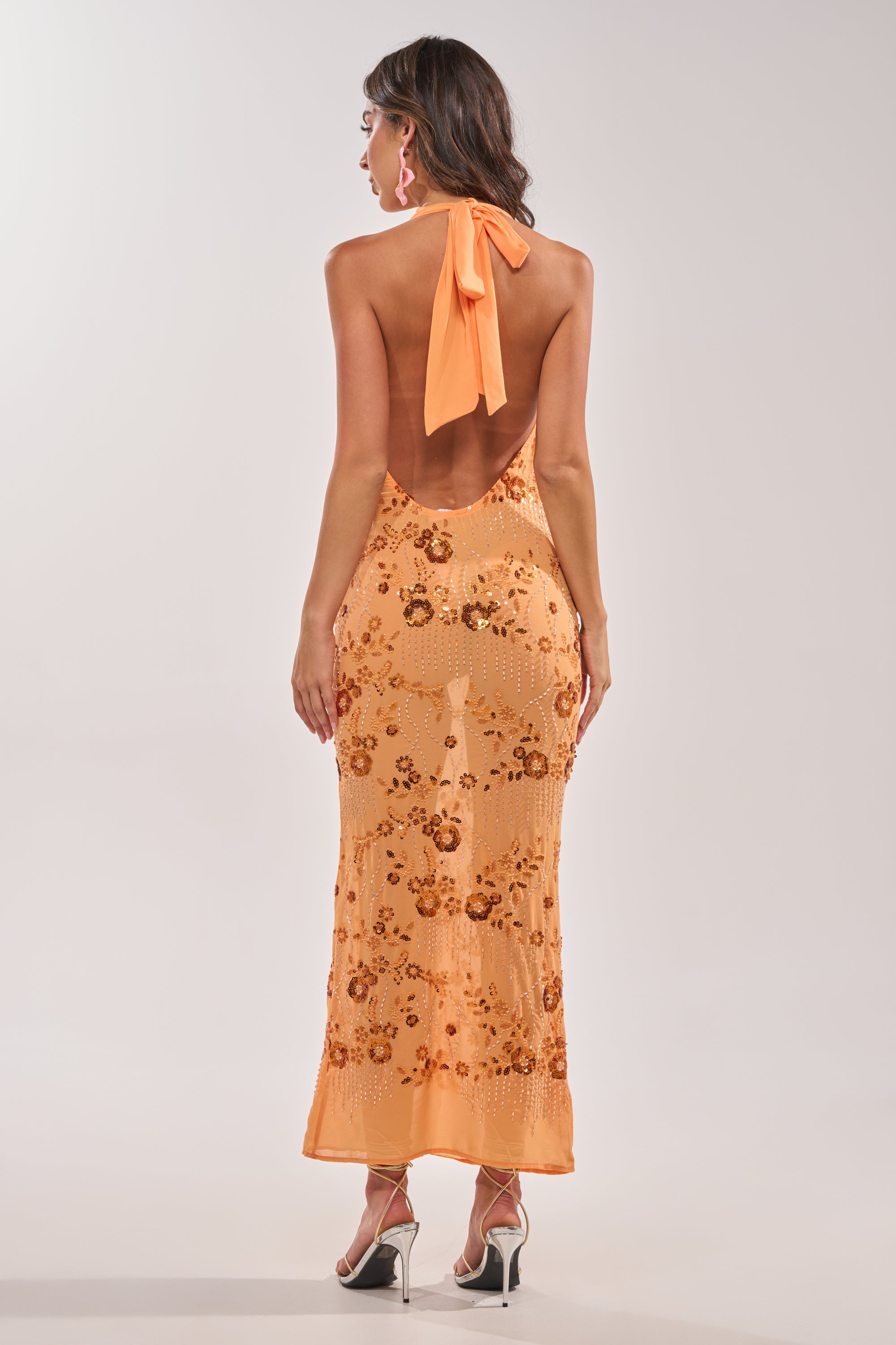 A woman stands facing away in the LOVE LETTERS EMBELLISHED MAXI DRESS IN ORANGE, featuring sequin floral patterns, an ankle-length backless halter design, and high heels against a plain light background. 5