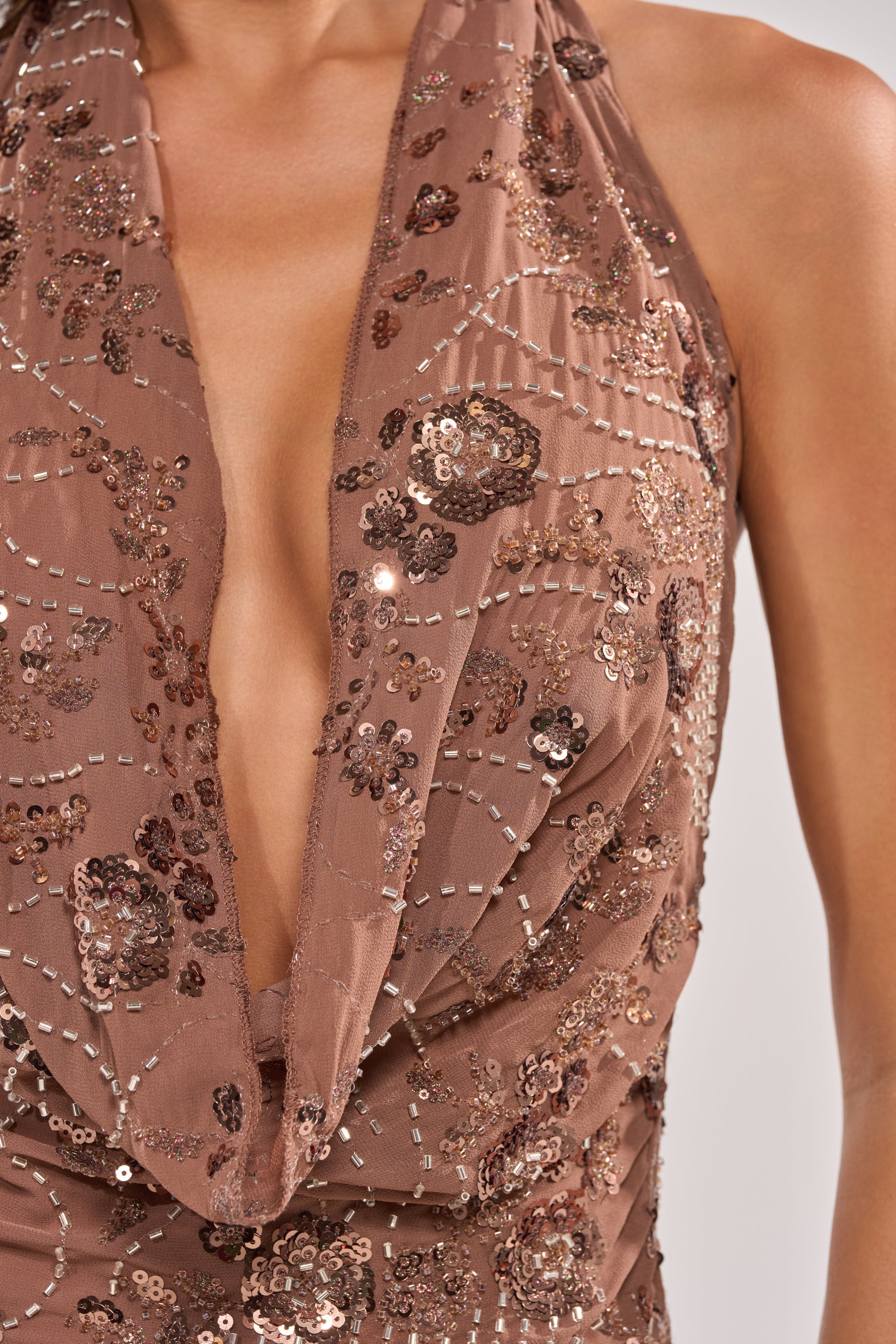 A close-up of a person wearing the LOVE LETTERS EMBELLISHED MAXI DRESS, a brown halter maxi with a deep V-neck and bronze sequin floral embellishments, set against a plain light background. 6