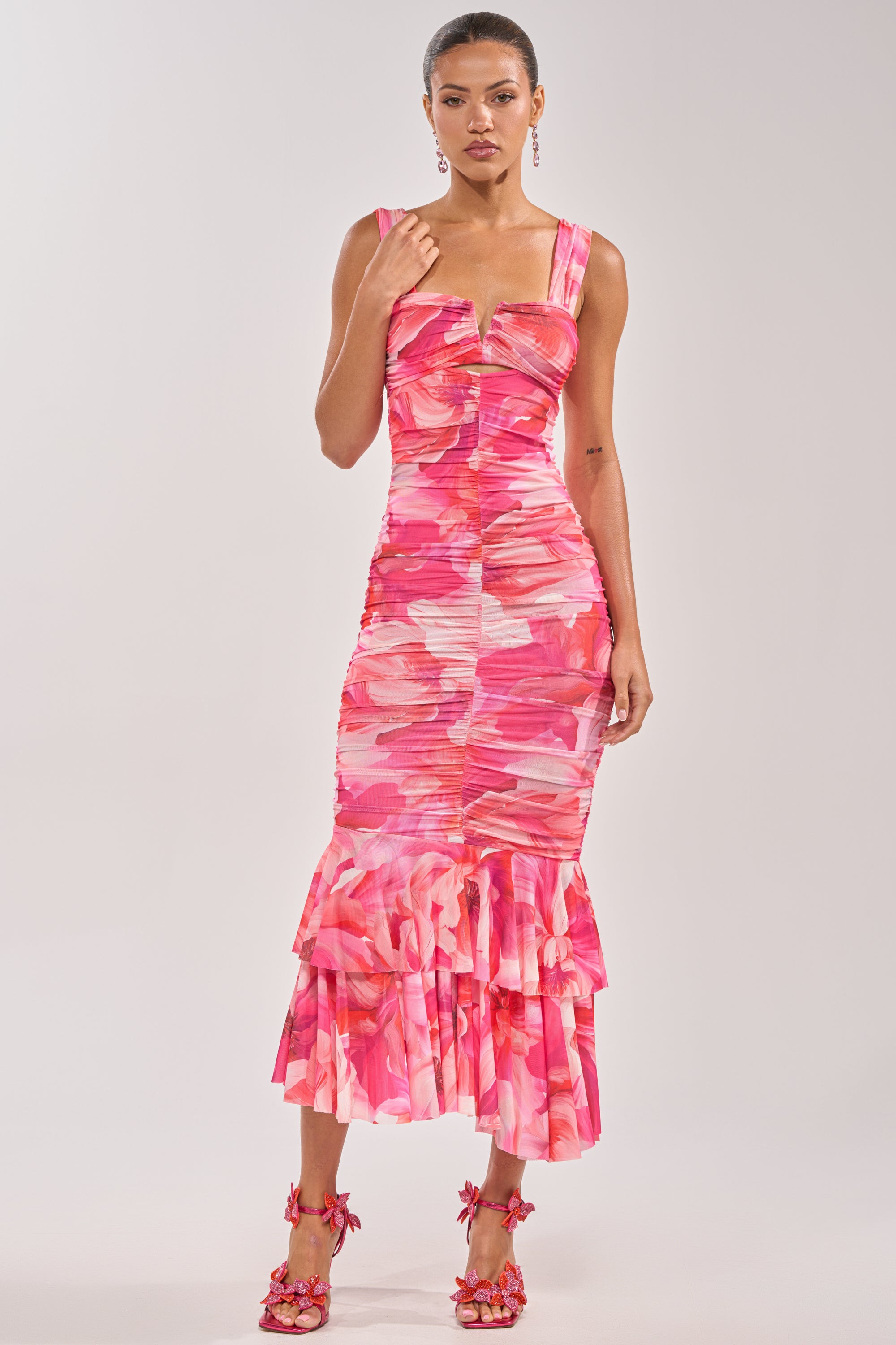 A woman poses against a plain light background wearing the BODY LANGUAGE MESH MAXI DRESS IN PINK, a fitted sleeveless maxi dress with an abstract floral print and ruffle hem, paired with matching floral heels. 1