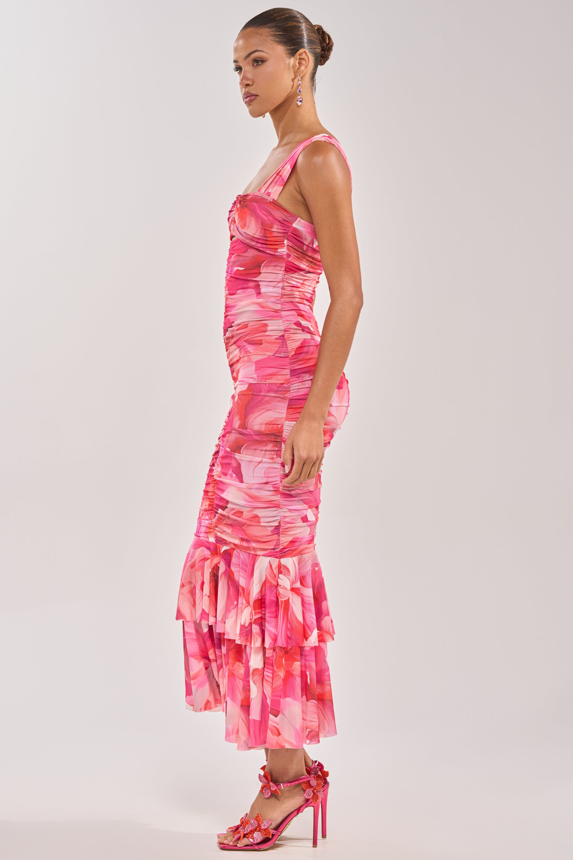 A woman stands in profile wearing the BODY LANGUAGE MESH MAXI DRESS IN PINK, a sleeveless, fitted abstract floral print dress with ruching and a mermaid hem. She pairs it with high-heeled sandals, a neat bun, and dangling earrings. 2
