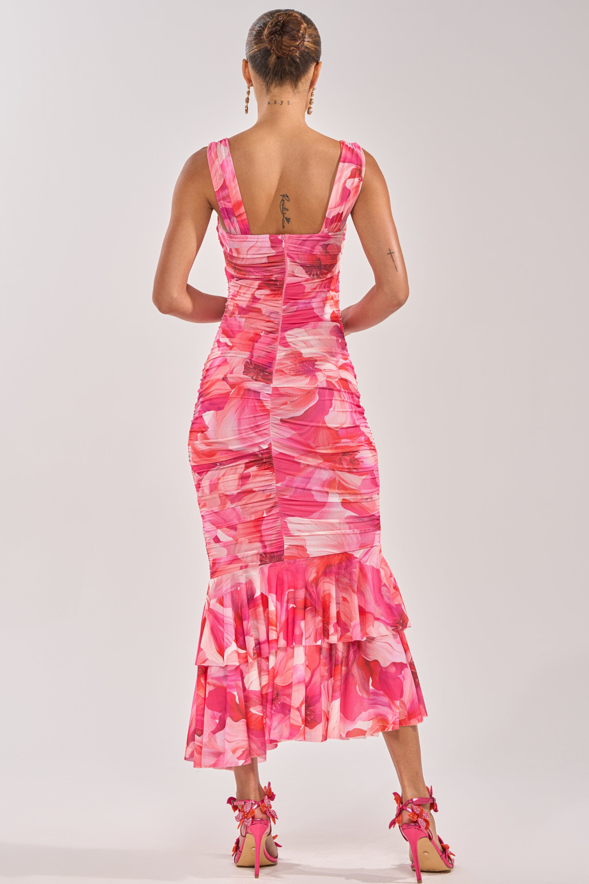 A woman faces away wearing the BODY LANGUAGE MESH MAXI DRESS IN PINK, featuring an abstract floral print and ruched mermaid hem, paired with matching heels. She has a neat bun and visible tattoos on her upper back and left arm. 3