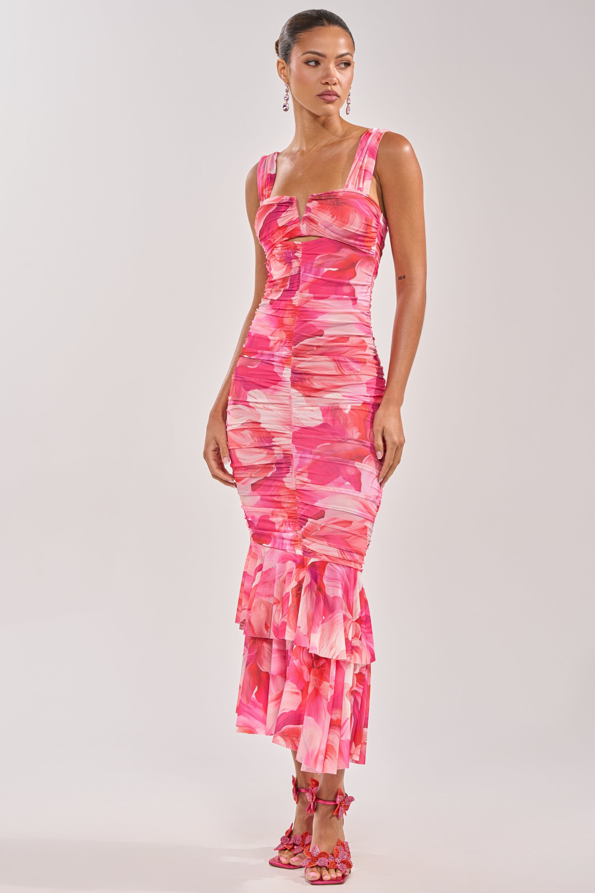 A woman wears the BODY LANGUAGE MESH MAXI DRESS IN PINK, featuring ruched details, pink and red abstract patterns, a ruffled hem, her hair pulled back, styled with matching strappy heels and dangling earrings. 4