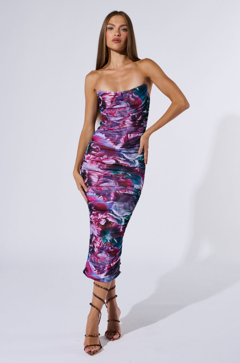 A woman with long hair stands confidently in a strapless, form-fitting FLORAL FANTASY RUCHED MIDI DRESS showcasing a vibrant abstract pattern in purple, blue, and pink. She is wearing strappy high-heeled sandals and poses against a plain white background.