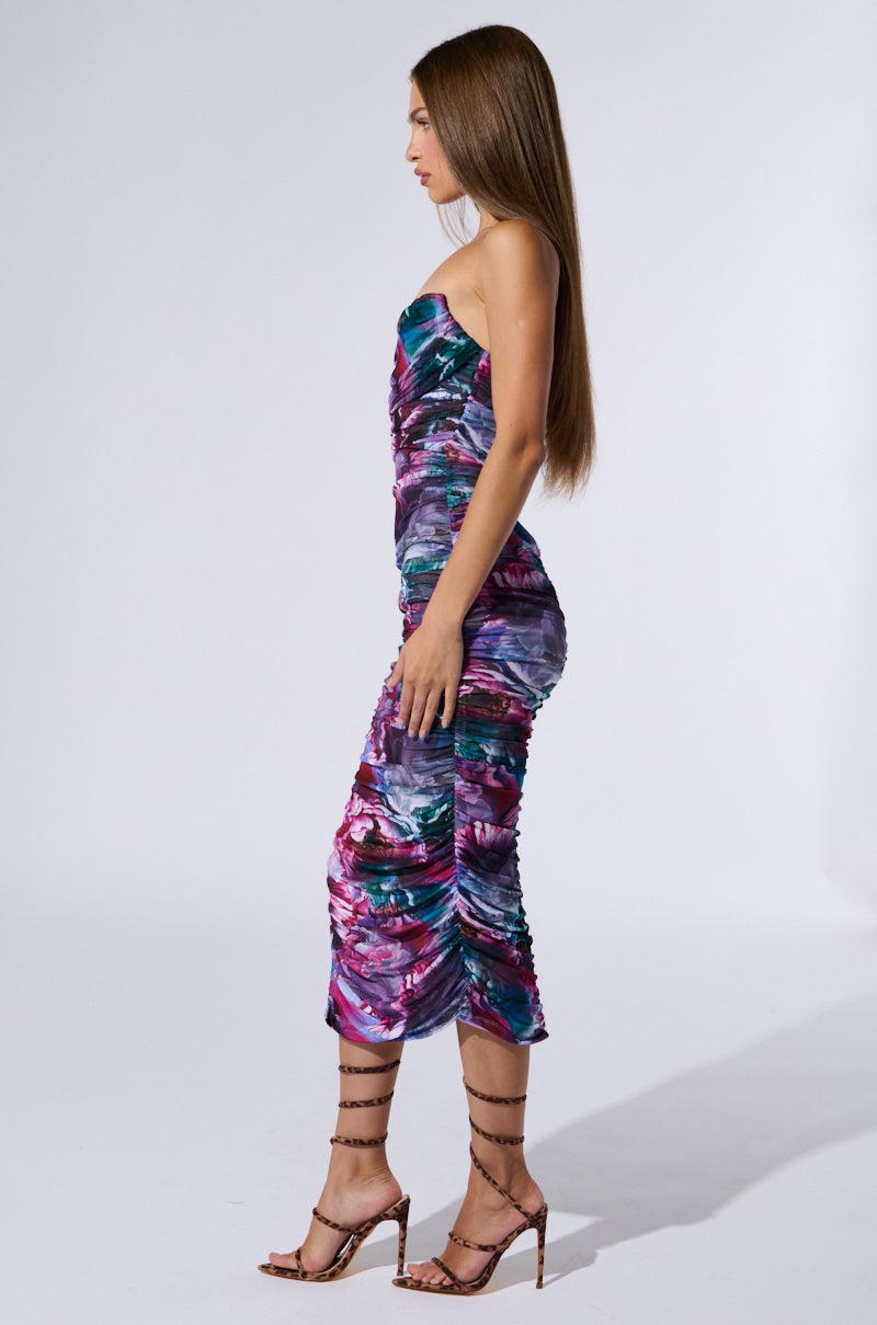 A woman with long hair stands in profile against a plain white background. She is wearing the FLORAL FANTASY RUCHED MIDI DRESS, a strapless design that captivates with its colorful pattern and bodycon fit, paired perfectly with high heels.