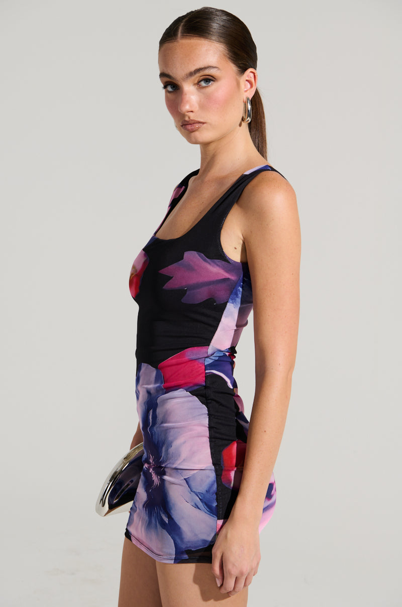 A woman in the NOT YOUR TOY FLORAL MESH MINI DRESS, featuring a fitted, sleeveless design with a vibrant floral pattern on a dark background, stands against a plain backdrop holding a small clutch. Her long, straight hair is pulled back and she accessorizes with hoop earrings. The bodycon silhouette of the dress is emphasized by its stretchy fabrication.
