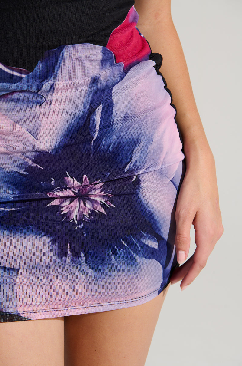 A person is posing in the NOT YOUR TOY FLORAL MESH MINI DRESS, featuring a colorful abstract print in blue, purple, pink, and black. The bodycon silhouette elegantly hugs their figure while the stretchy mesh fabric ensures comfort. One hand rests gracefully alongside the vibrant skirt.