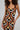 A person with curly hair wears the ZIGGY MIDI DRESS IN ORANGE, featuring bold, abstract Art Deco print patterns in orange, black, and gold. The curve-hugging silhouette is accentuated by a wide gold bracelet on their upper arm against a plain grey background.