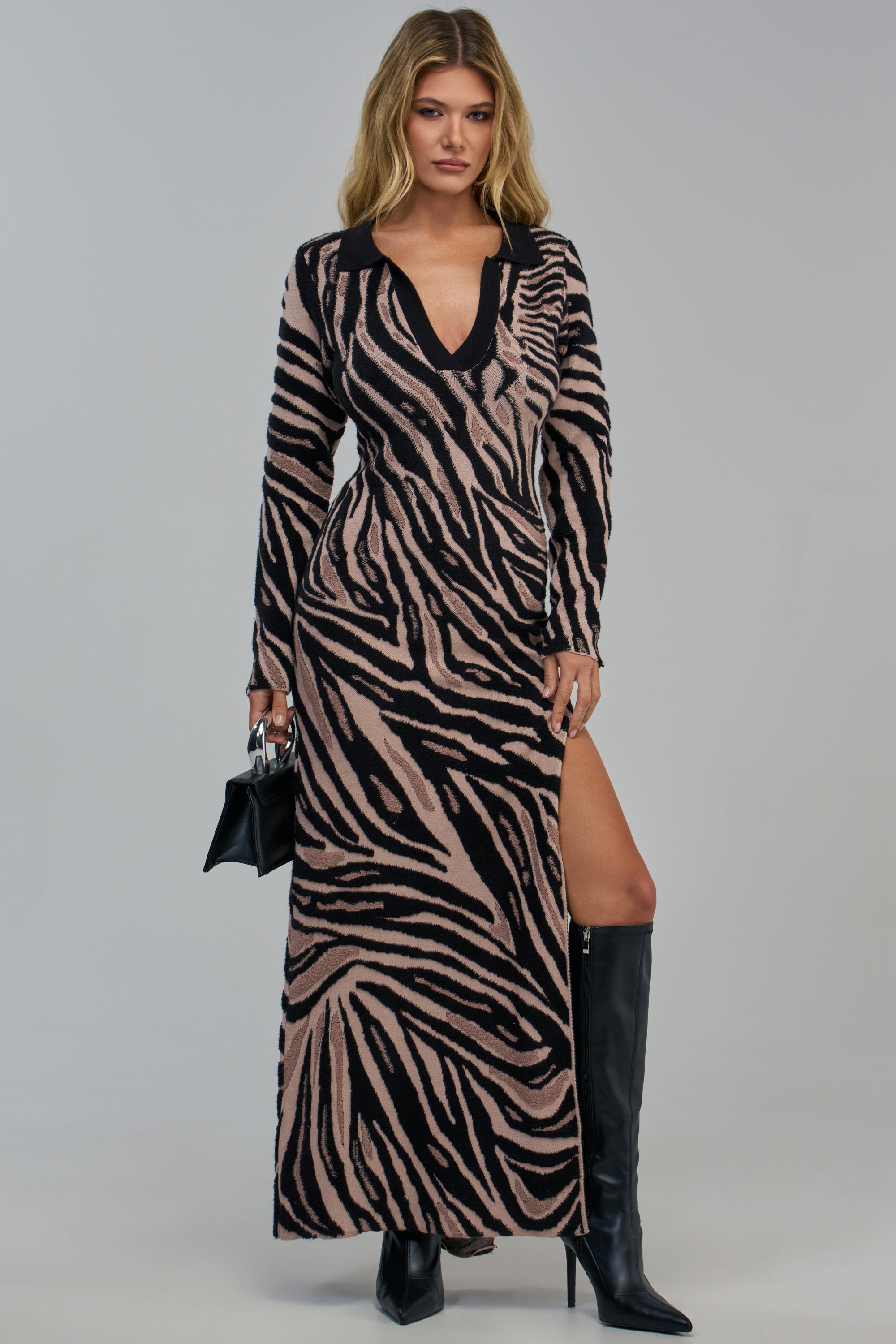 A woman models the HARD TO FORGET COLLARED SWEATER DRESS IN BLACK MULTI, a long-sleeved tiger print maxi dress with a deep V-neck and high side slit, paired with black knee-high boots and a small black handbag.