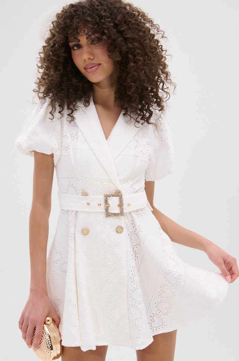 A woman with curly hair models the TRUE DELIGHT BELTED MINI DRESS featuring puff sleeves and lace detailing. She holds a gold clutch and stands against a plain white background, smiling slightly. 1