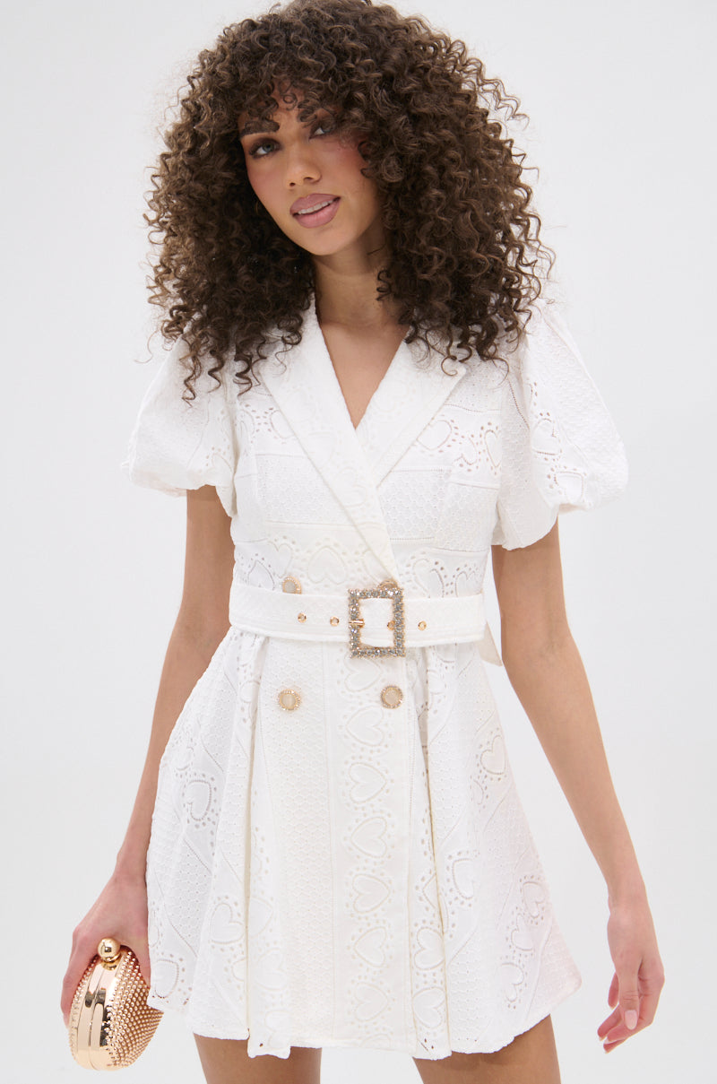 A woman with curly hair smiles slightly while wearing the TRUE DELIGHT BELTED MINI DRESS, which features puff sleeves and textured patterns. She holds a small metallic clutch and stands against a plain light background. 2