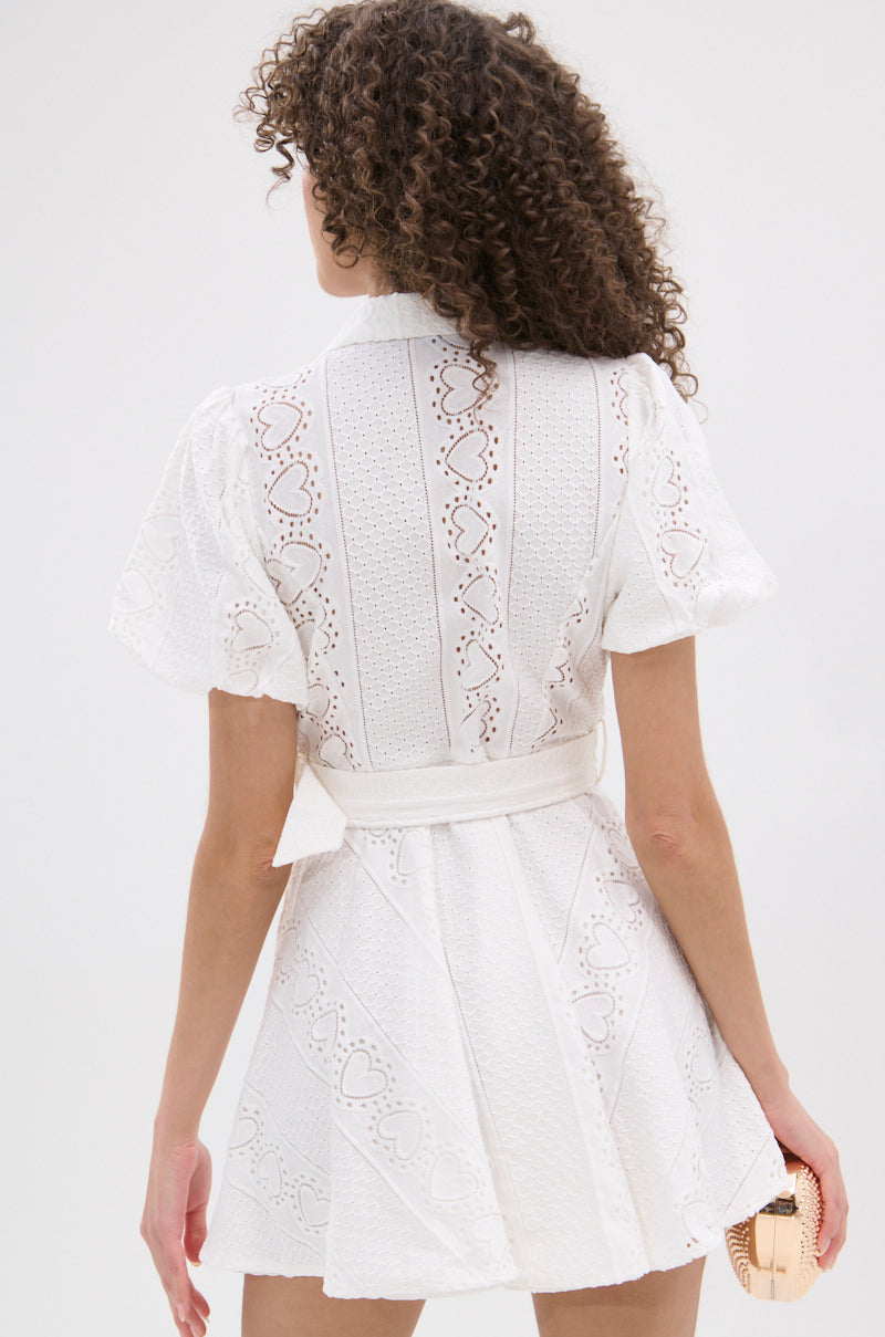 A woman with curly hair is shown from the back wearing the TRUE DELIGHT BELTED MINI DRESS, a white fit-and-flare mini dress with a heart-shaped eyelet pattern. She holds a small light-colored handbag in her left hand. 4