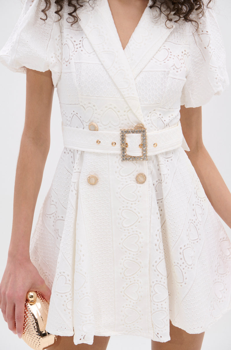 A woman wears the TRUE DELIGHT BELTED MINI DRESS in white lace with puff sleeves, rhinestone buttons, and a jeweled-buckle belt. She holds a shiny gold clutch in her left hand; only her torso and part of her arm are visible. 7
