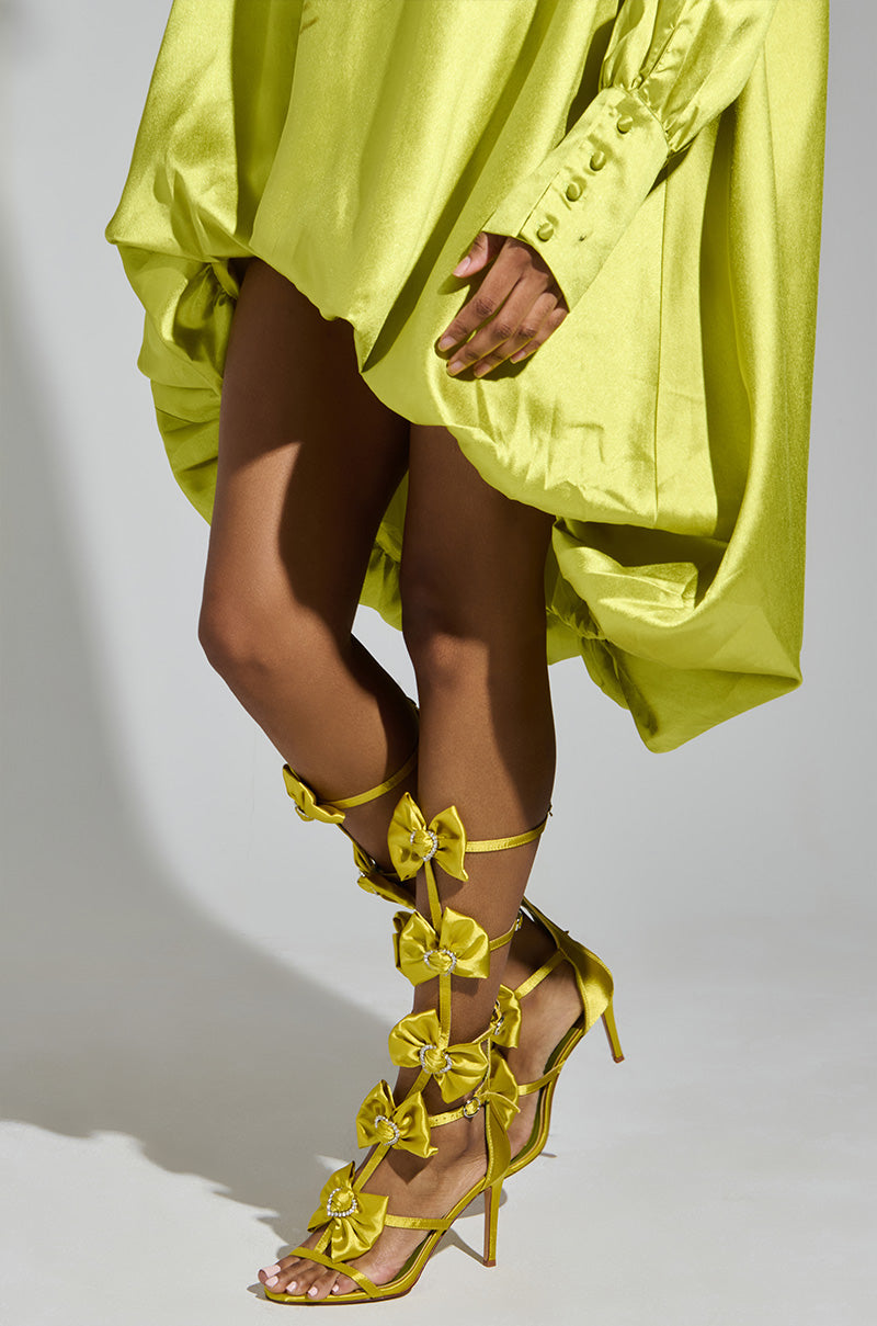 Someone is wearing a flowing lime green dress with AZALEA WANG AERIN LIME BOW EMBELLISHED GLADIATOR STILETTO SANDALS featuring comfort cushion insoles and large bows. The button details on the sleeves add flair to this vibrant look against a plain background, accentuating the unique shoe design. 2