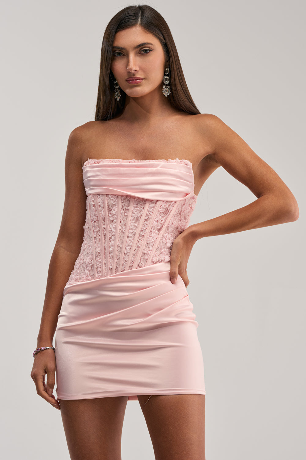 A woman poses against a plain background in the RADIANT RUSH LACE CORSET MINI DRESS, a strapless light pink mini dress with lace and pleated details, fitted bodice, and dangling earrings.