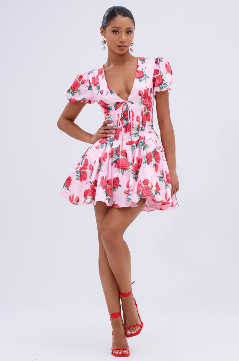 A woman models the WILL YOU TAKE THIS ROSE SATIN MINI DRESS, featuring pink floral print, puff sleeves, and a deep V-neckline. She pairs it with red high-heeled sandals and poses confidently against a plain background.