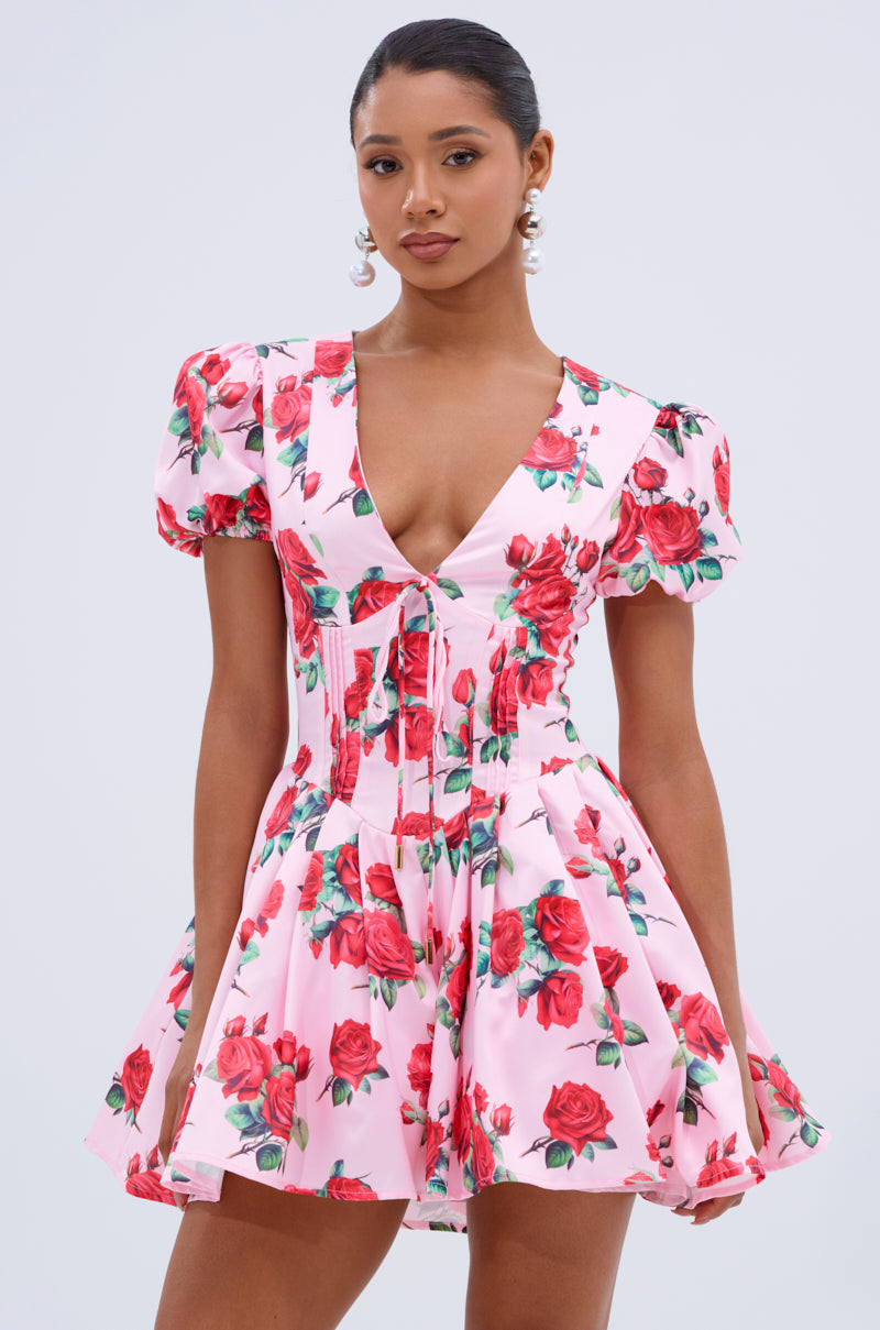 A woman poses against a plain background in the WILL YOU TAKE THIS ROSE SATIN MINI DRESS, a pink floral mini with red rose pattern, deep V neckline, flared skirt, and short sleeves for a charming, elegant style.