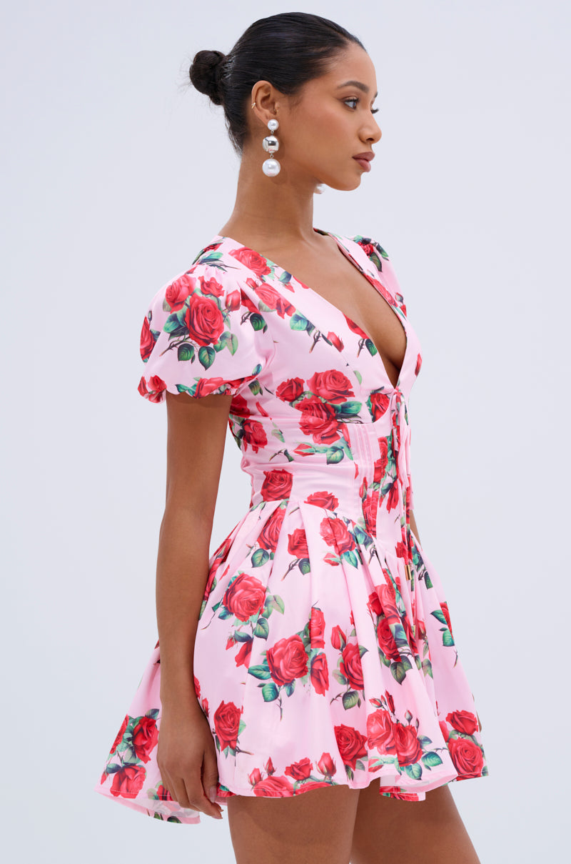 A woman wears the WILL YOU TAKE THIS ROSE SATIN MINI DRESS, featuring pink floral print, puff sleeves, and a deep V-neckline. She accessorizes with pearl drop earrings and wears her hair in a neat bun, standing in profile against a plain background.