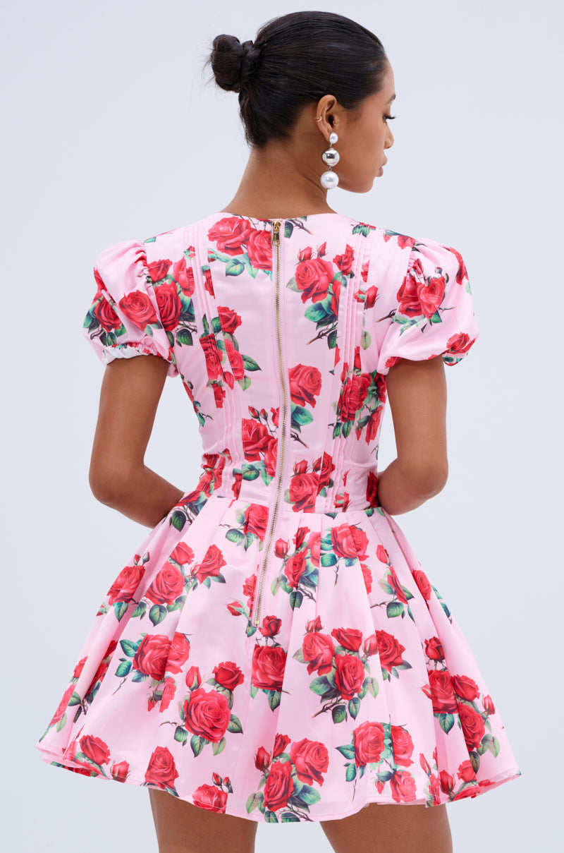 A woman with her hair in a bun models the WILL YOU TAKE THIS ROSE SATIN MINI DRESS, showcasing its puff sleeves, red rose print, and back zipper. She faces away from the camera, accessorized with large dangling pearl earrings.