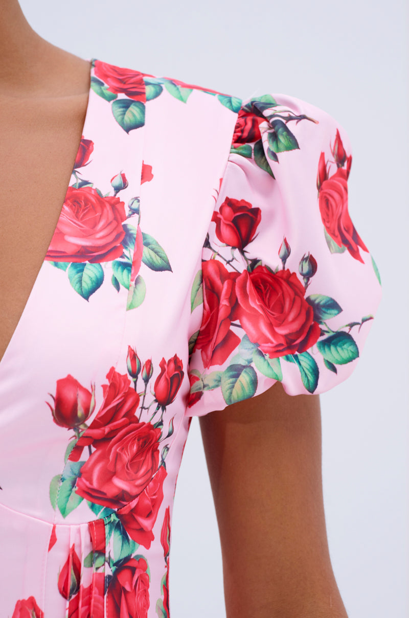 A close-up of a person wearing the WILL YOU TAKE THIS ROSE SATIN MINI DRESS, a light pink mini dress with puff sleeves, deep V-neckline, and vibrant red rose prints with green leaves. The background is plain and light.