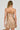 A person with braided hair is dressed in the GOLDEN GIRL CHAINED UP MINI DRESS, embellished with gold sequins and fringe. They carry a matching purse and display several tattoos on their back and arms. The bodycon silhouette contrasts beautifully against the white background, accentuating the outfit's elegance.
