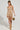 Dressed in the GOLDEN GIRL CHAINED UP MINI DRESS, a woman poses confidently, showcasing its bodycon silhouette that features gold sparkles and fringe details. She holds a small gold bag and wears strappy heels, with the plain background allowing her glamorous elegance to shine through.
