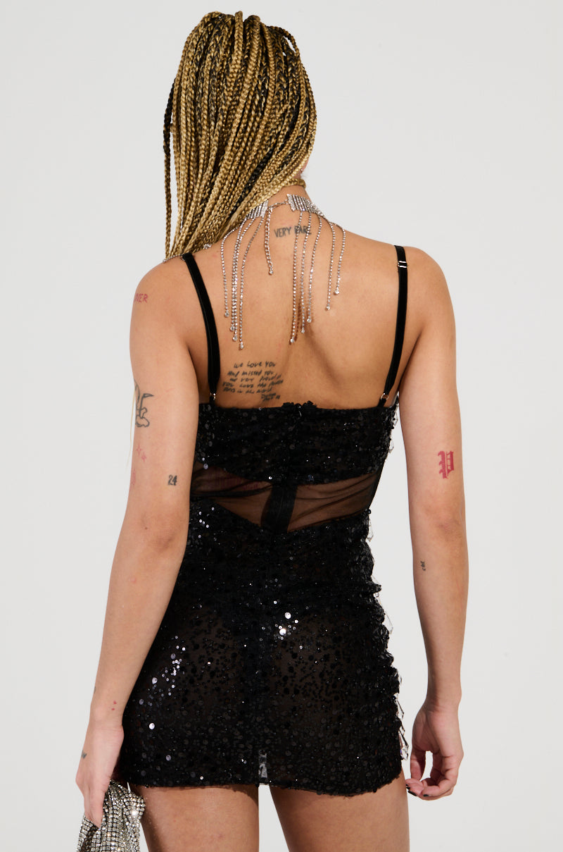 A person with braided hair stands with their back to the camera, wearing a JULIETTE CRYSTAL EMBELLISHED MINI DRESS that sparkles with sequin and bead embellishments. Tattoos adorn their back and arms, complemented by a necklace featuring chains that drape elegantly across their shoulders.