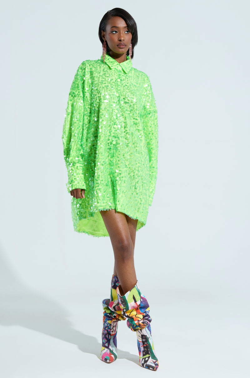 A person poses with confidence in a vivid green VERONICA SEQUIN COLLARED SHIRT DRESS, complemented by colorful, patterned high boots. The plain light blue backdrop accentuates the striking attire embellished with translucent sequin discs.